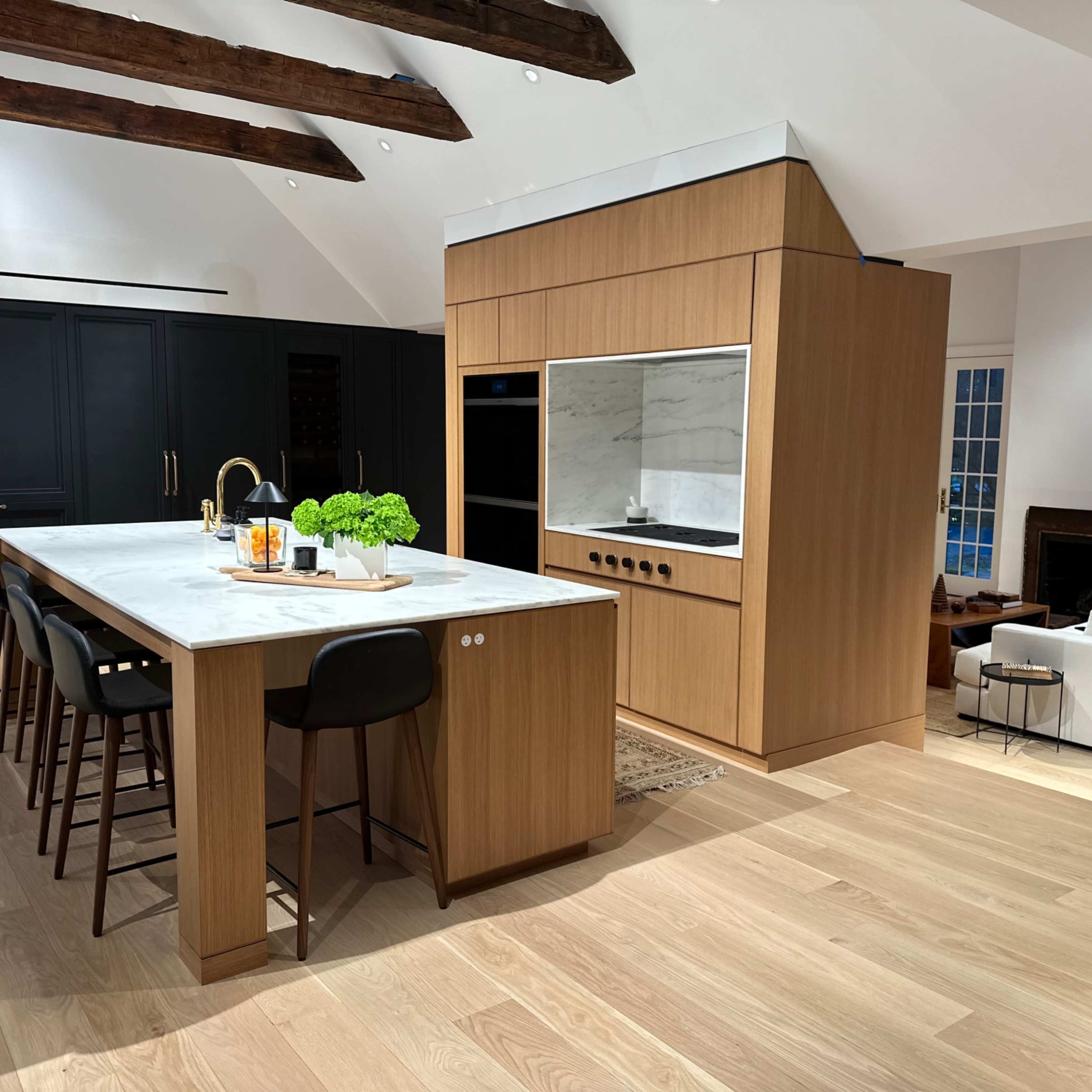 A modern kitchen features a large marble island with bar seating, wooden cabinetry, and a black accent wall, all set in an open living space.