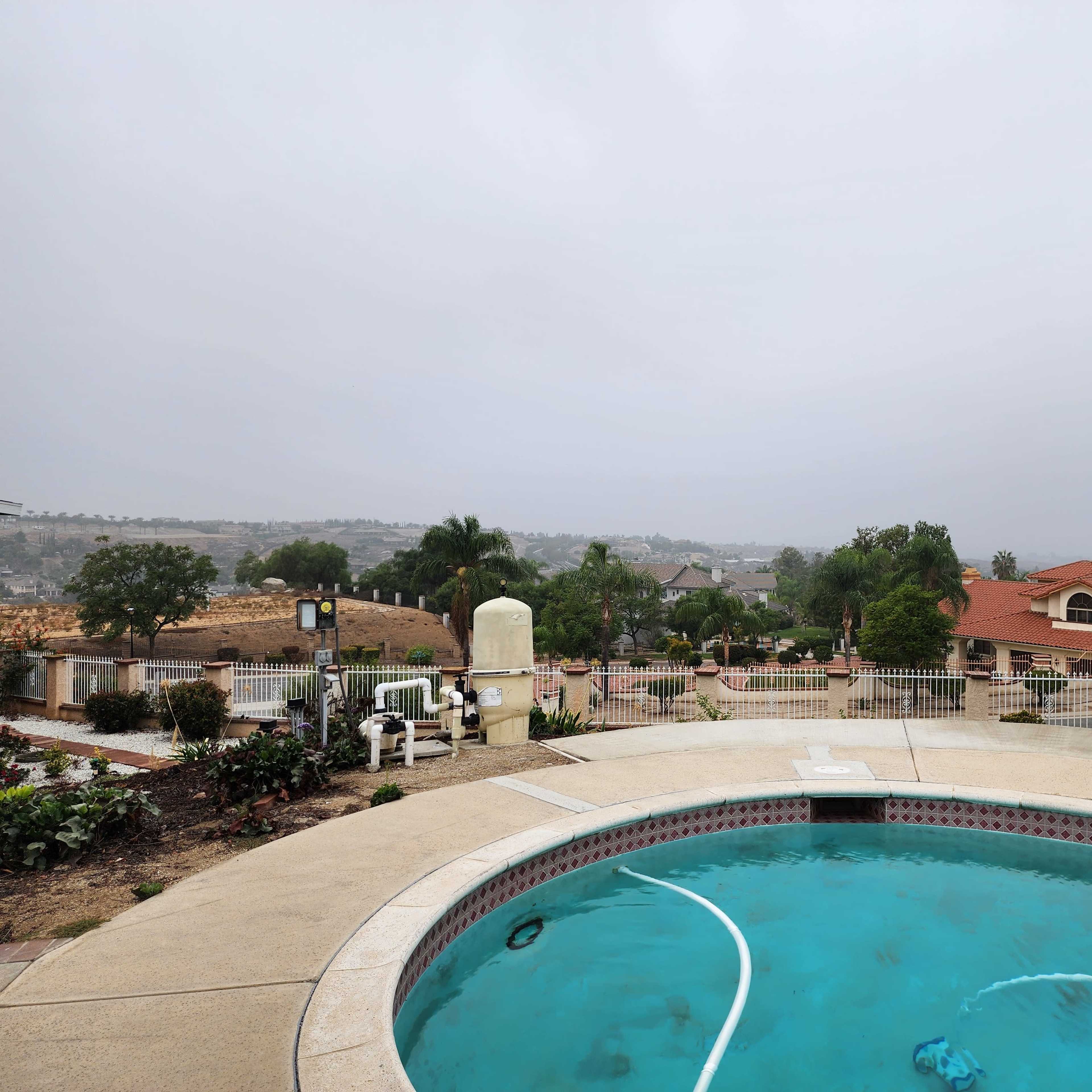 A circular swimming pool with a spa sits in an outdoor area surrounded by greenery and residential buildings under a cloudy sky.