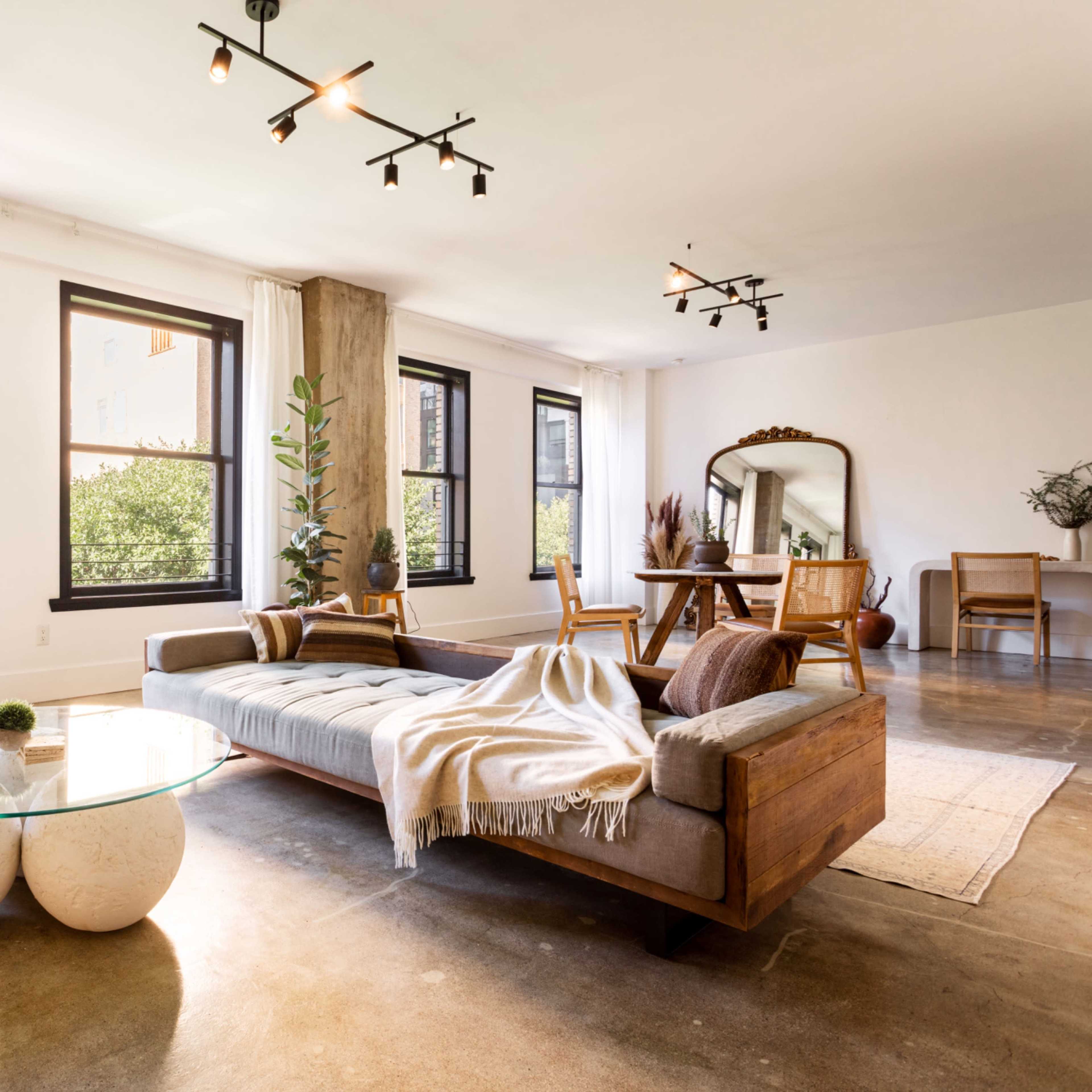 A modern living room with large windows, a wooden sofa, a circular glass coffee table, and a dining area featuring a wooden table and chairs.