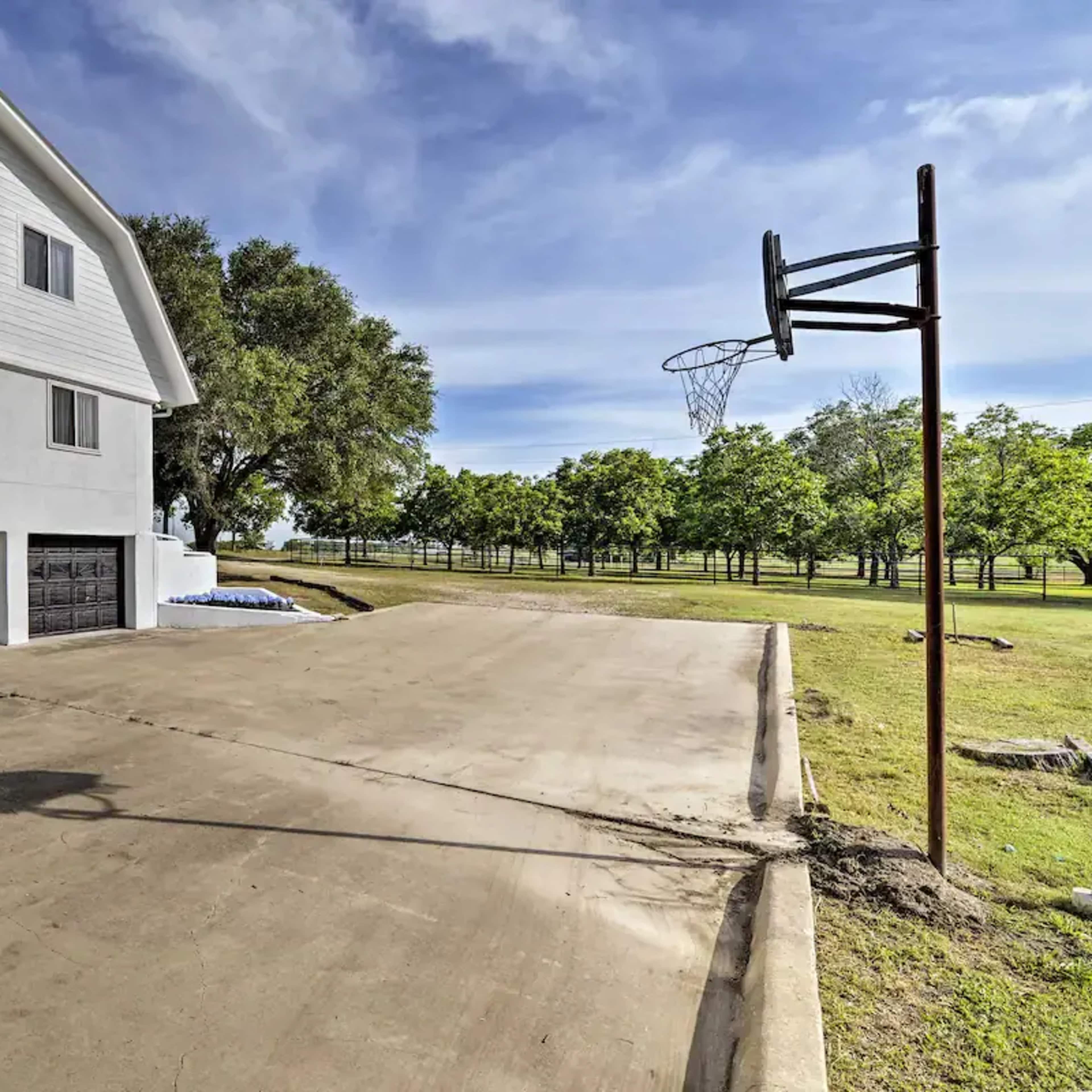 A white house with a large driveway and a basketball hoop stands next to a grassy area lined with trees.