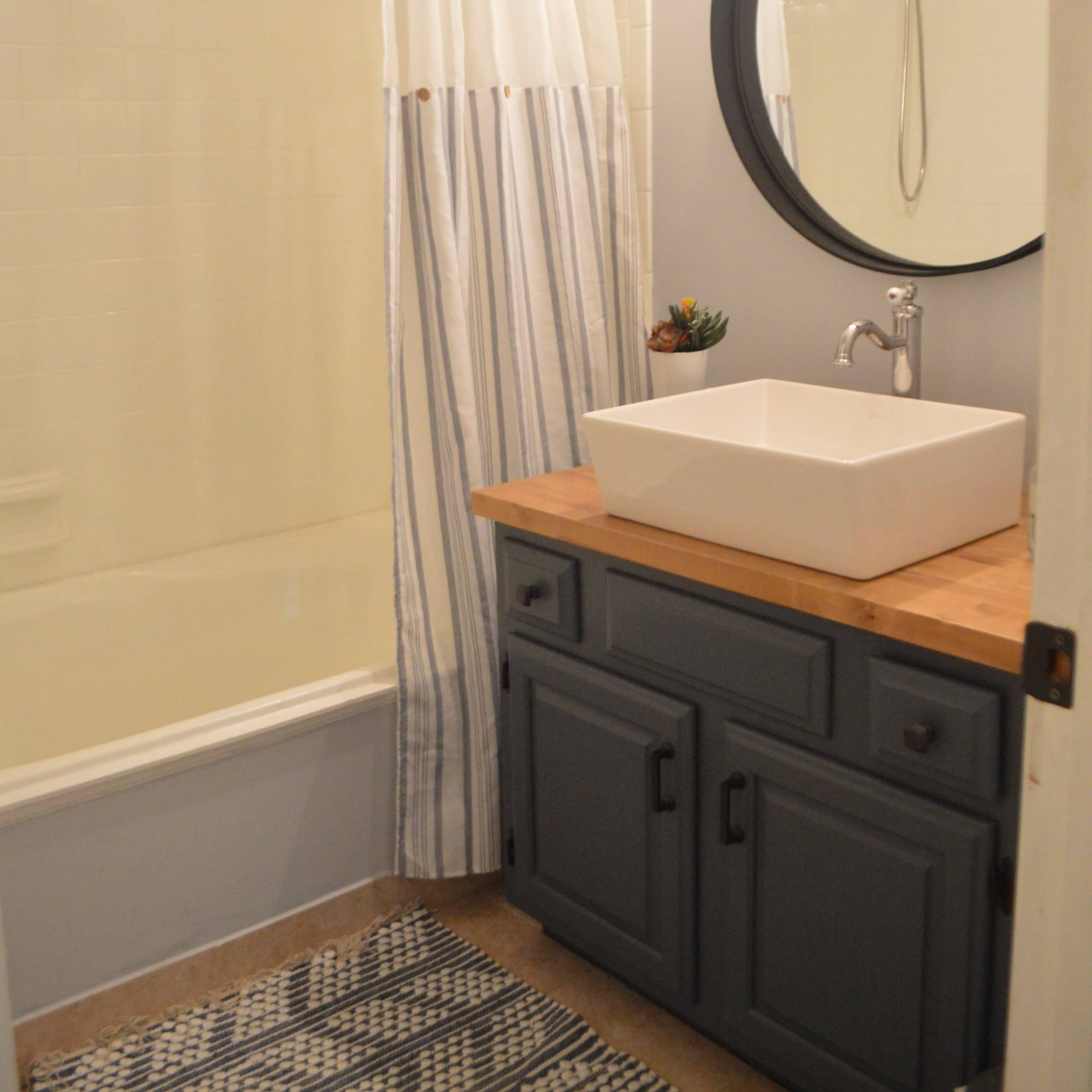 A bathroom featuring a bathtub with a shower curtain, a circular mirror, a rectangular sink on a wooden countertop, and a small potted plant.