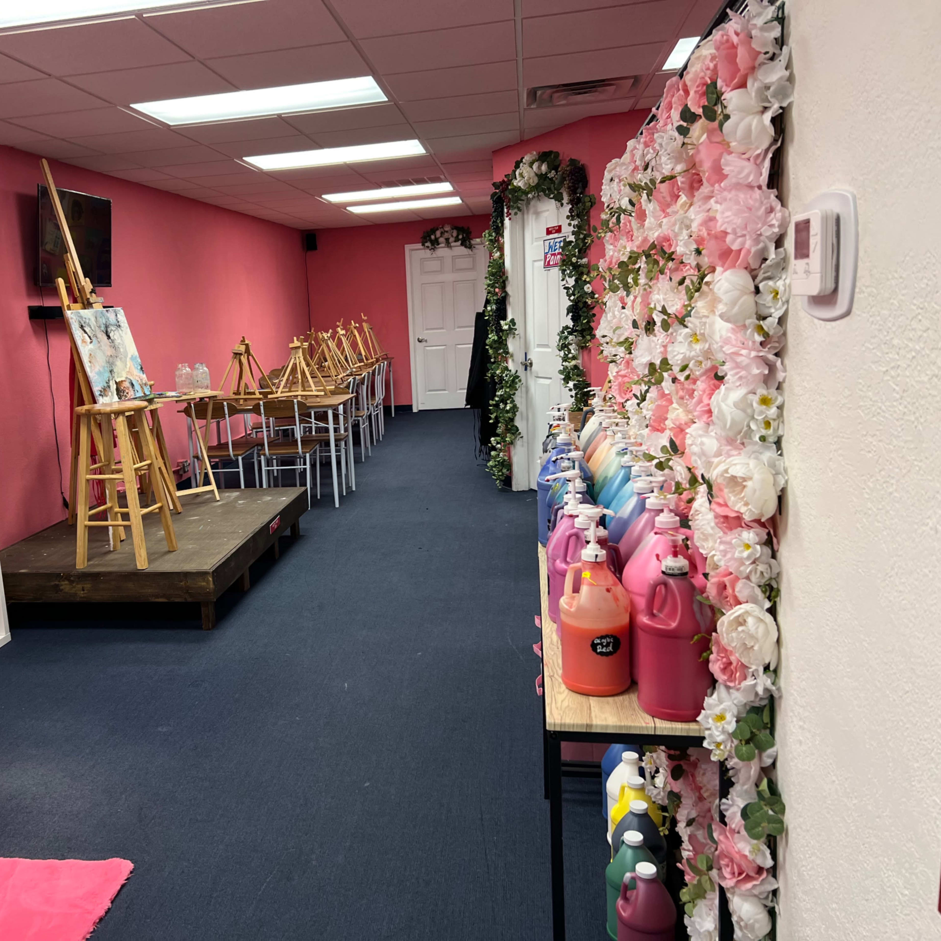 A brightly colored art studio with pink walls, a small stage for painting, easels set up, and neatly arranged bottles of paint along one side.
