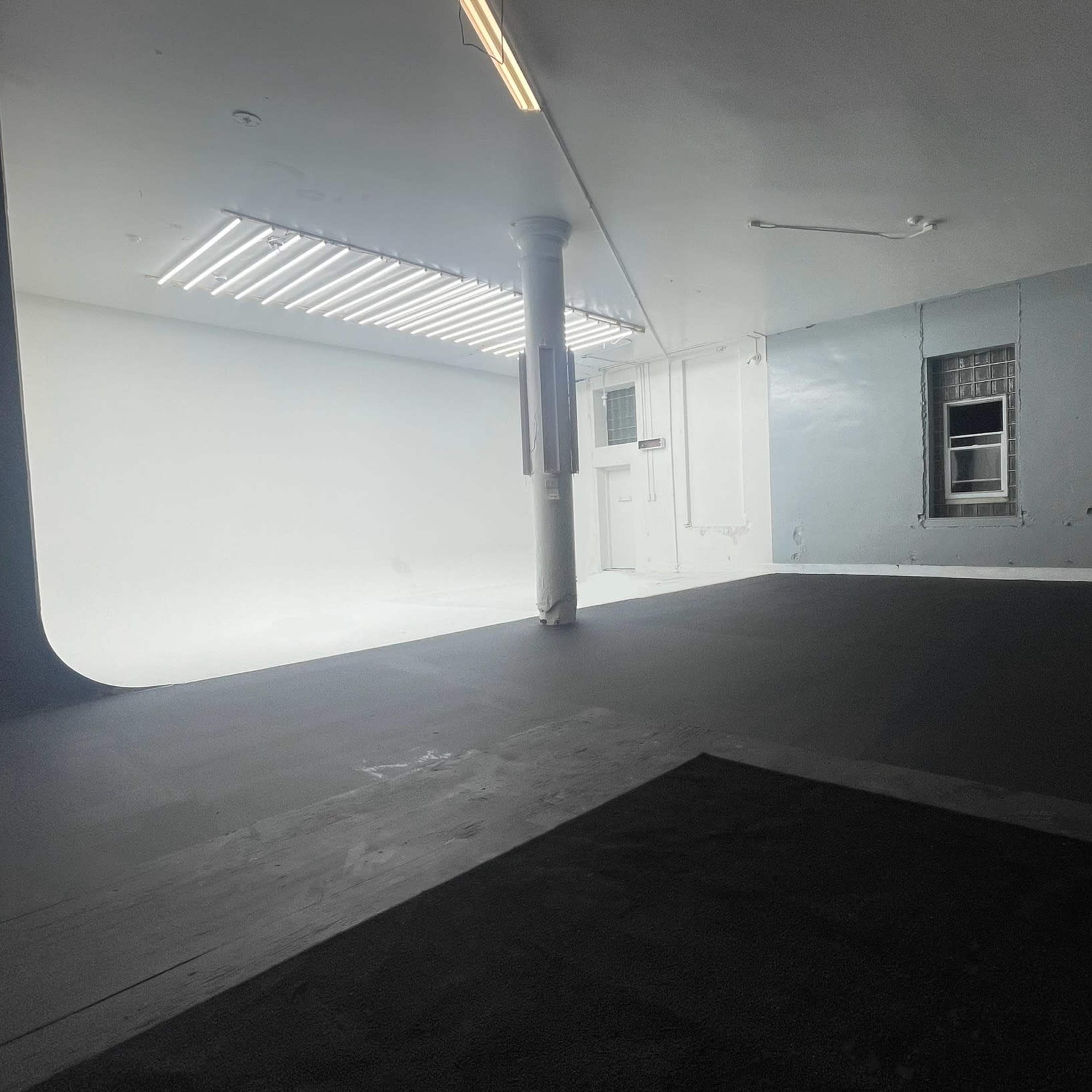 A spacious studio with black flooring, white walls, and a large open area illuminated by fluorescent lights.