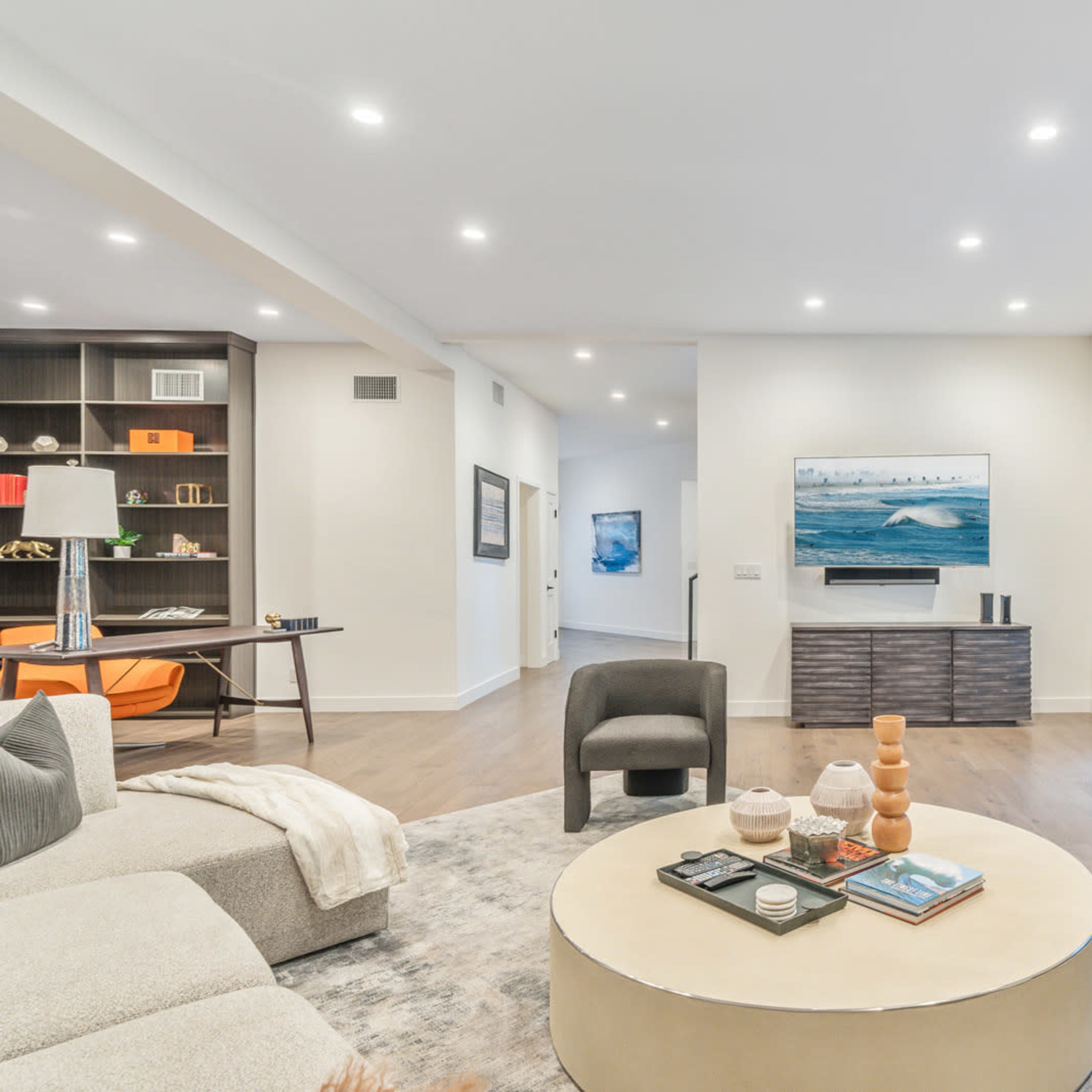 A modern, open-concept living room with a circular coffee table, a sectional sofa, and a television mounted on the wall, adjacent to large sliding glass doors leading to an outdoor area.