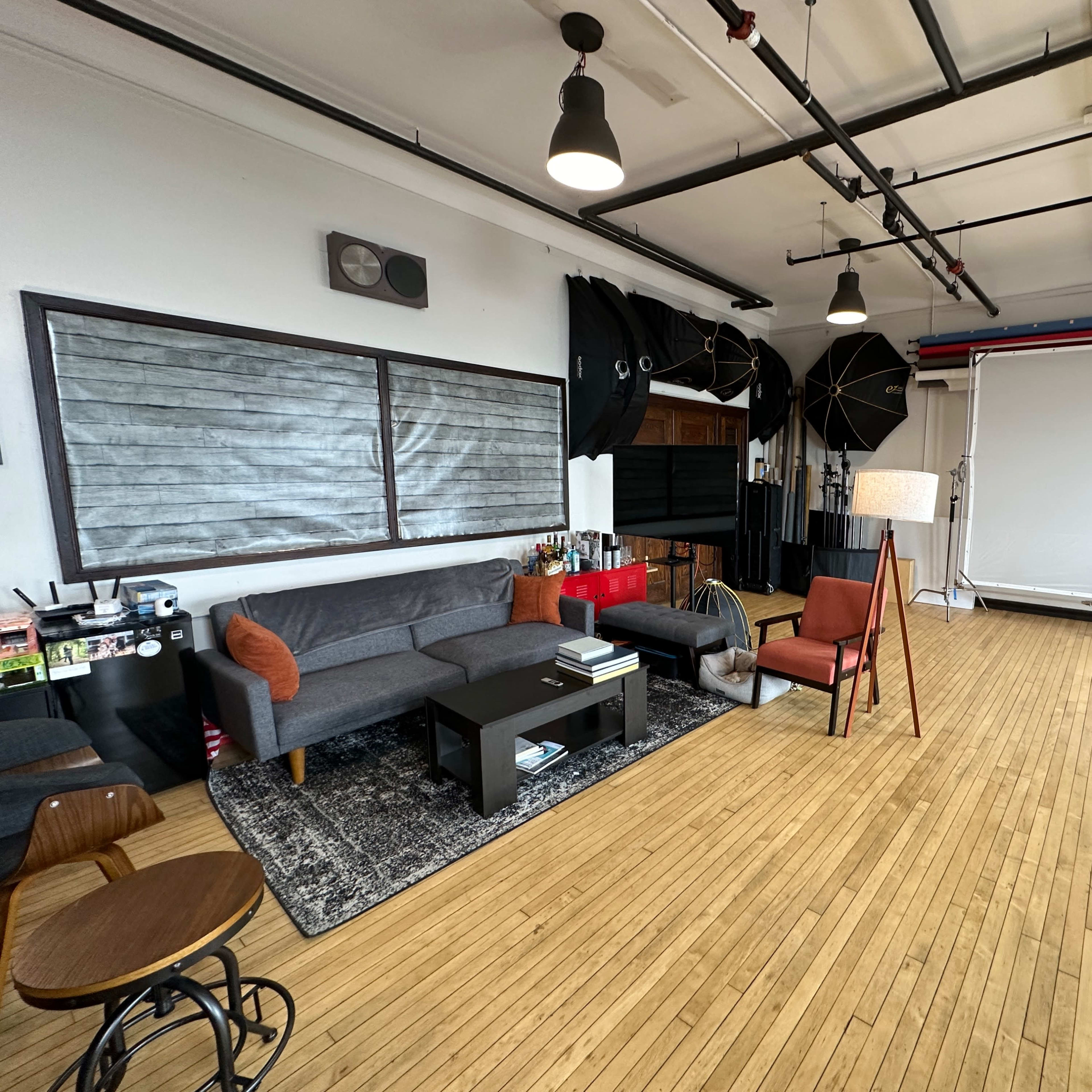 A spacious studio with a gray sofa, a brown chair, a black coffee table, and various photography equipment positioned around a large backdrop.