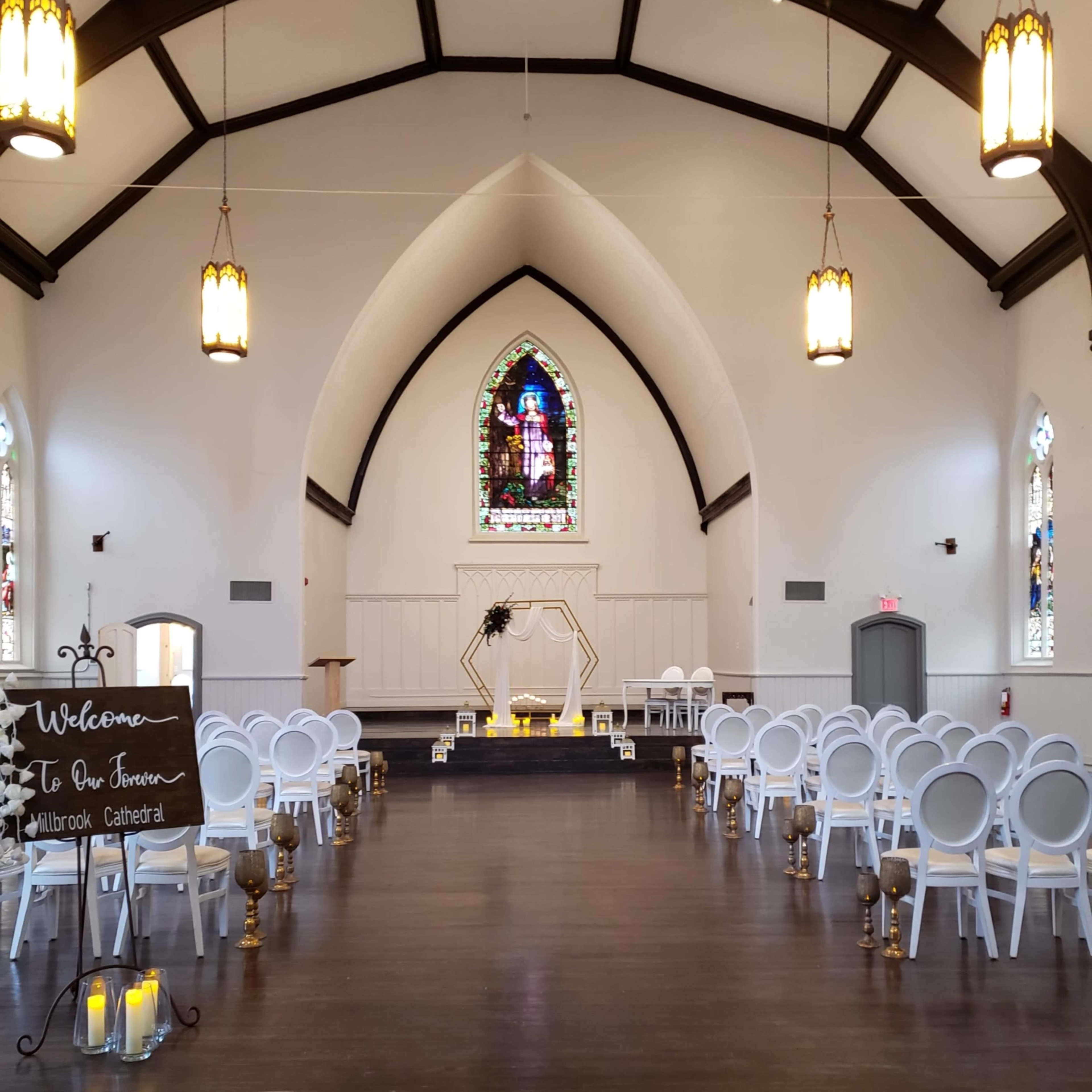 The interior of a church features rows of white chairs facing a decorative altar and a stained glass window at the far end.