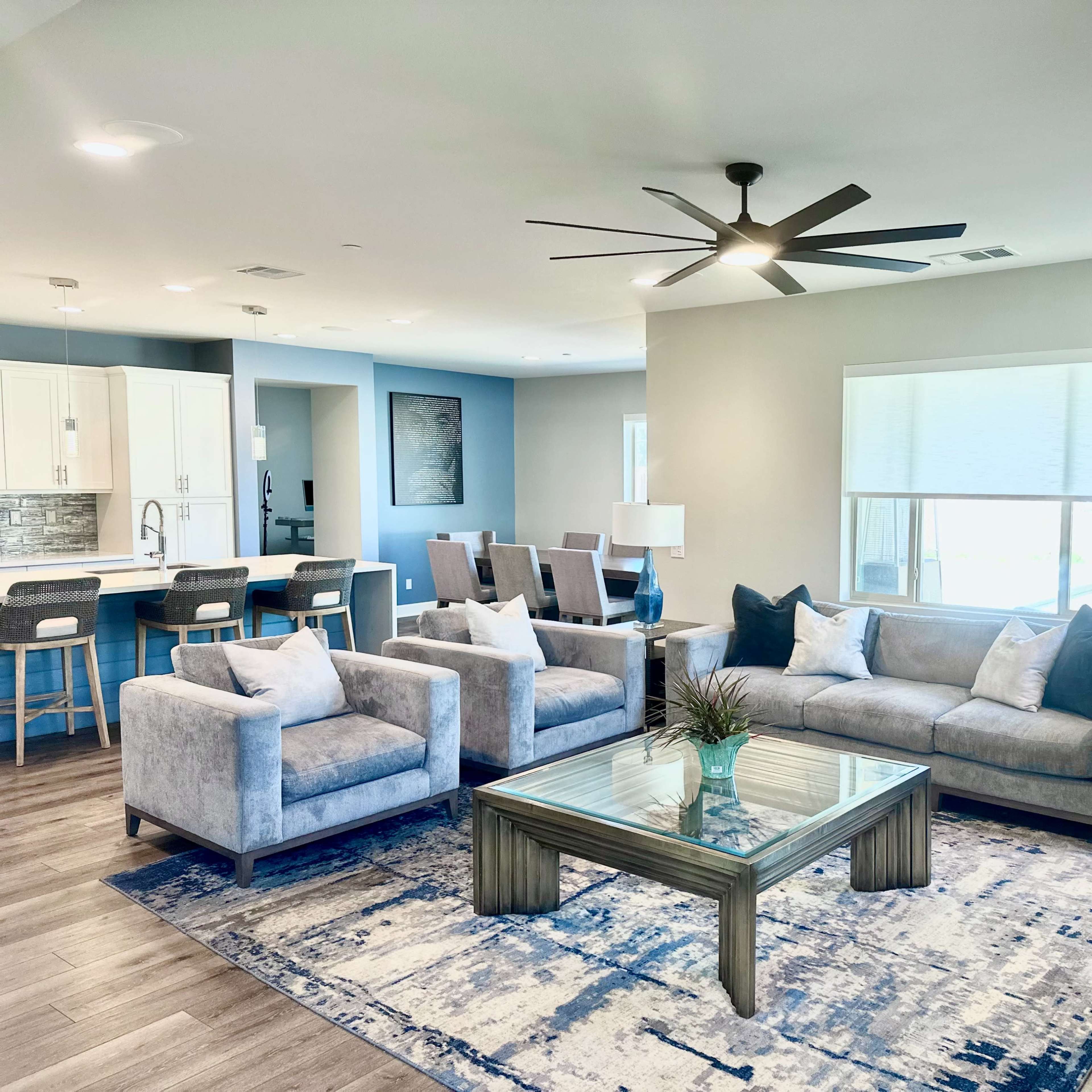 A modern living room with a gray sofa, two armchairs, a glass coffee table, and a large window, complemented by a kitchen area in the background.