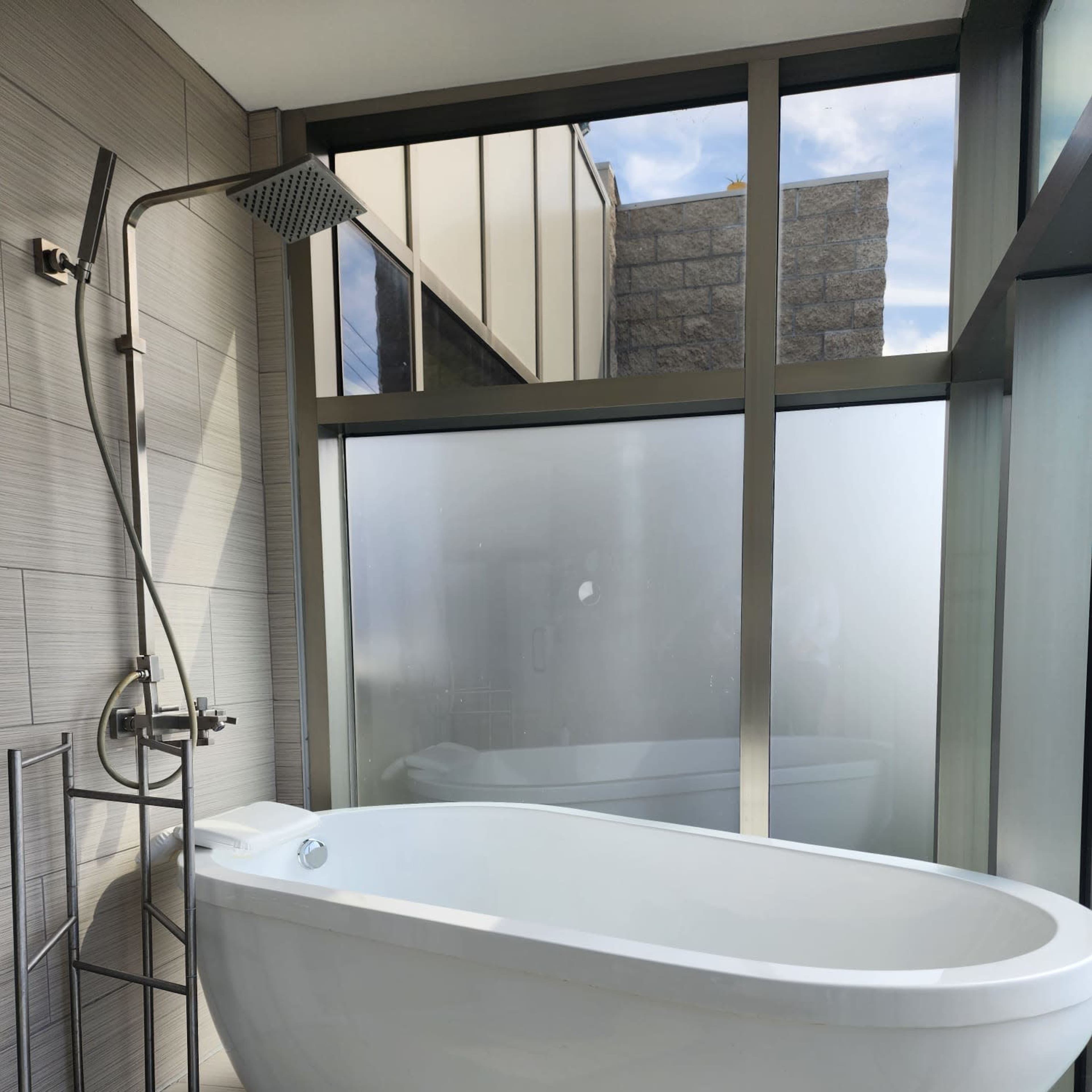 A modern bathroom features a freestanding white bathtub beside a glass wall with natural light streaming in.