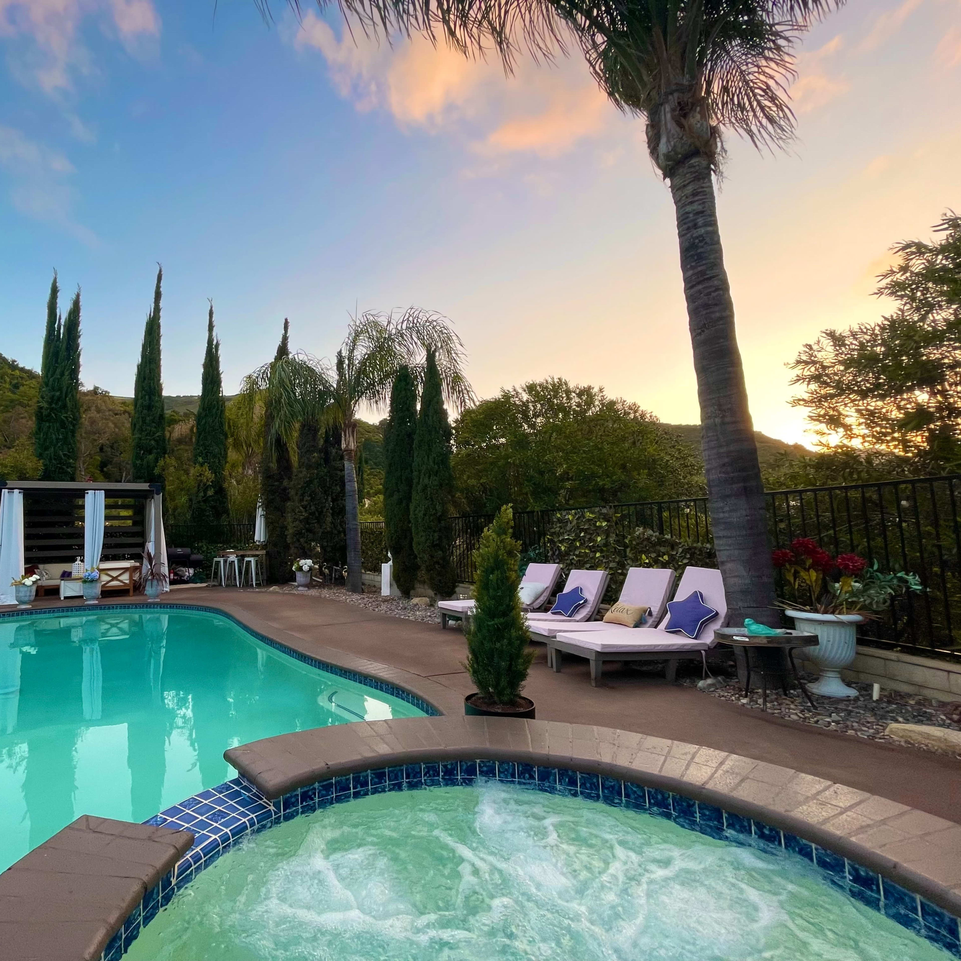 The scene features a swimming pool and spa surrounded by palm trees, lounge chairs, and a backdrop of lush hills under a colorful sky at sunset.