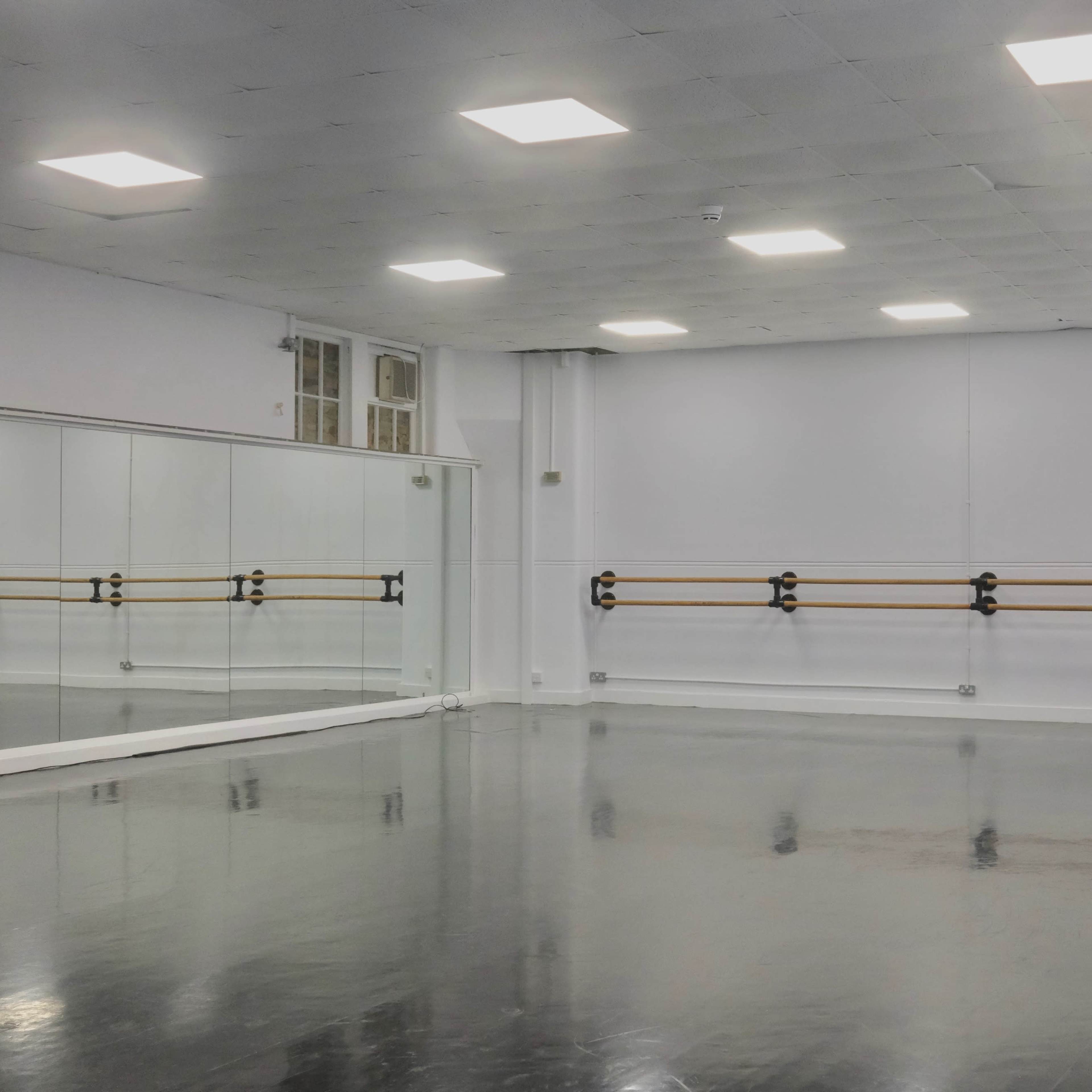 A spacious, empty dance studio with a wooden barre along the mirrored wall and a polished floor illuminated by overhead lights.