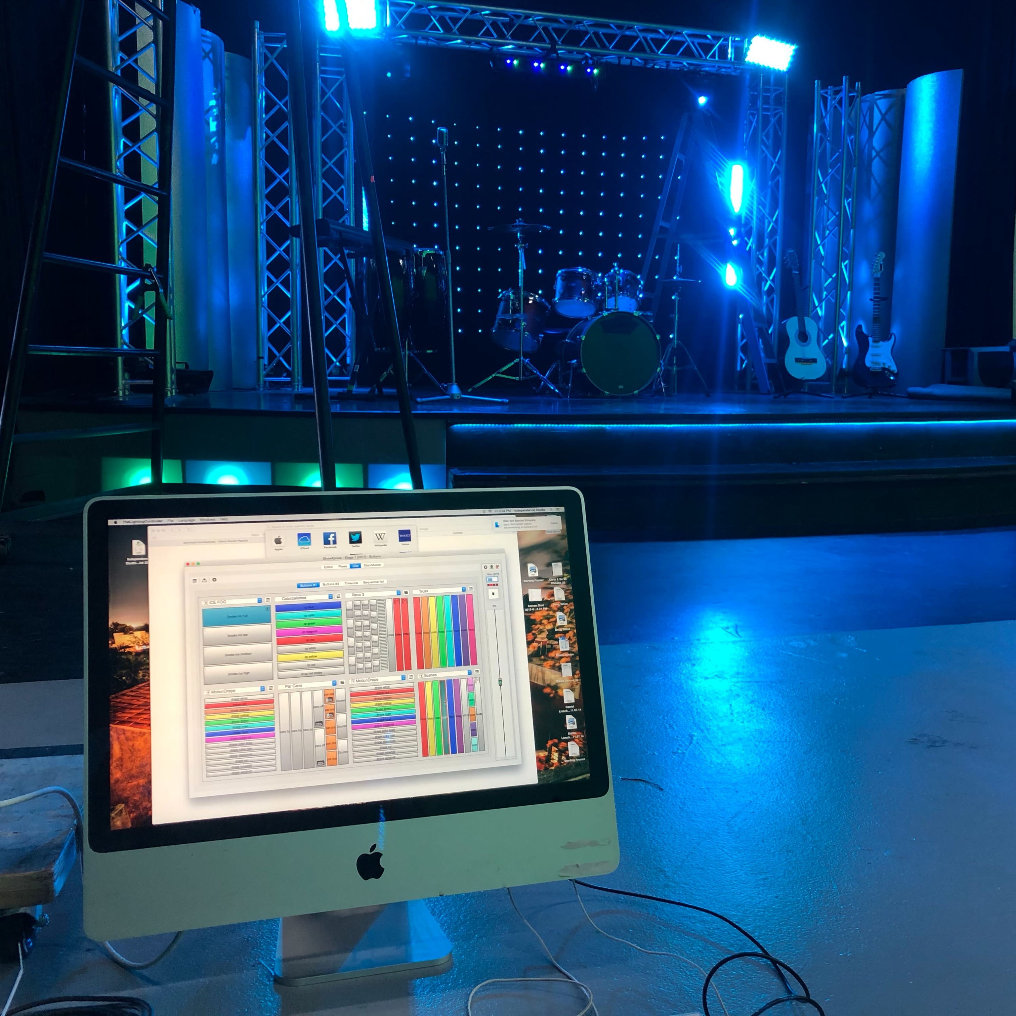 A computer displays software with a colorful interface in the foreground, while a stage set for a performance, featuring instruments and lighting, is visible in the background.