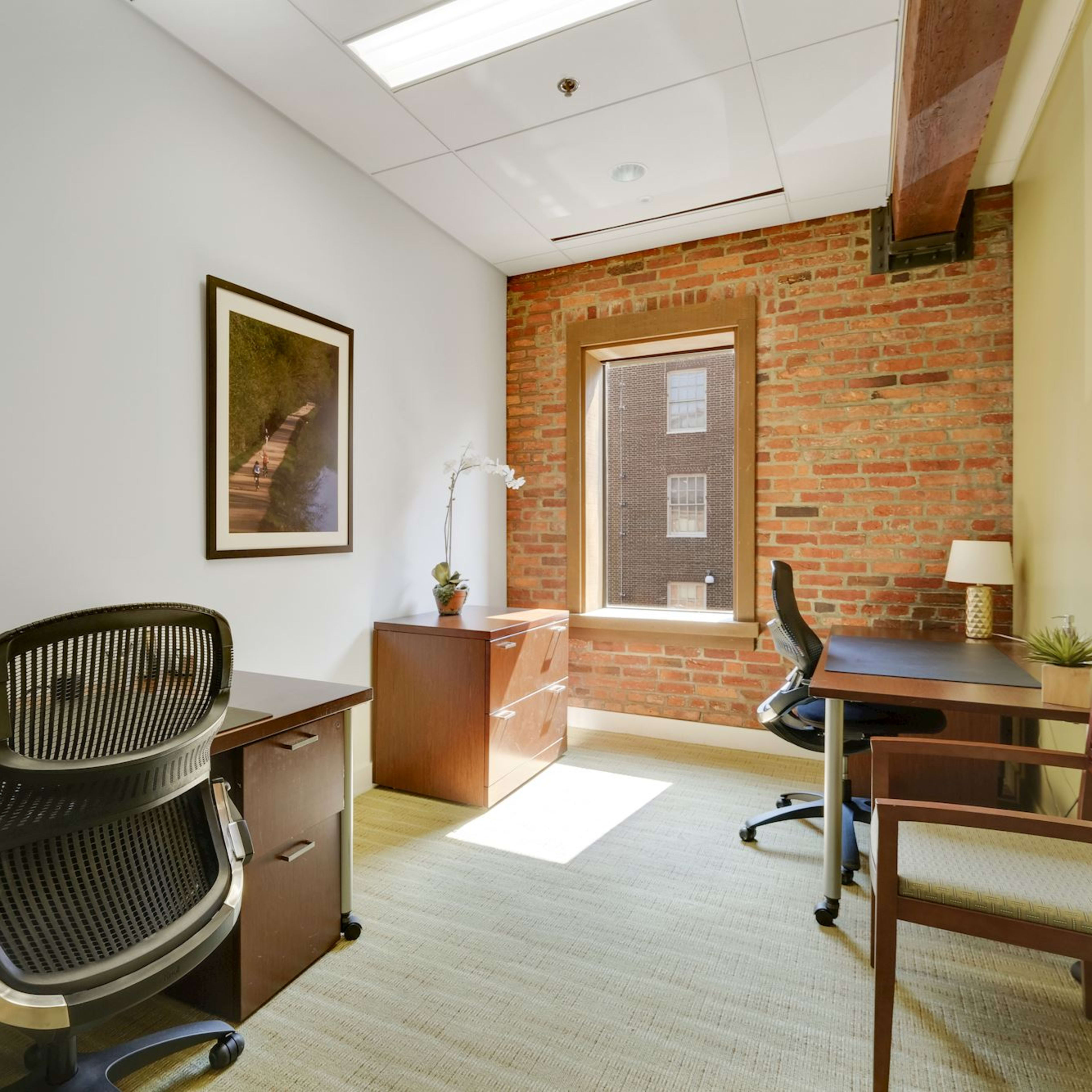 A small office space featuring two desks, chairs, a window with a view of brick buildings, and a potted plant on one desk.