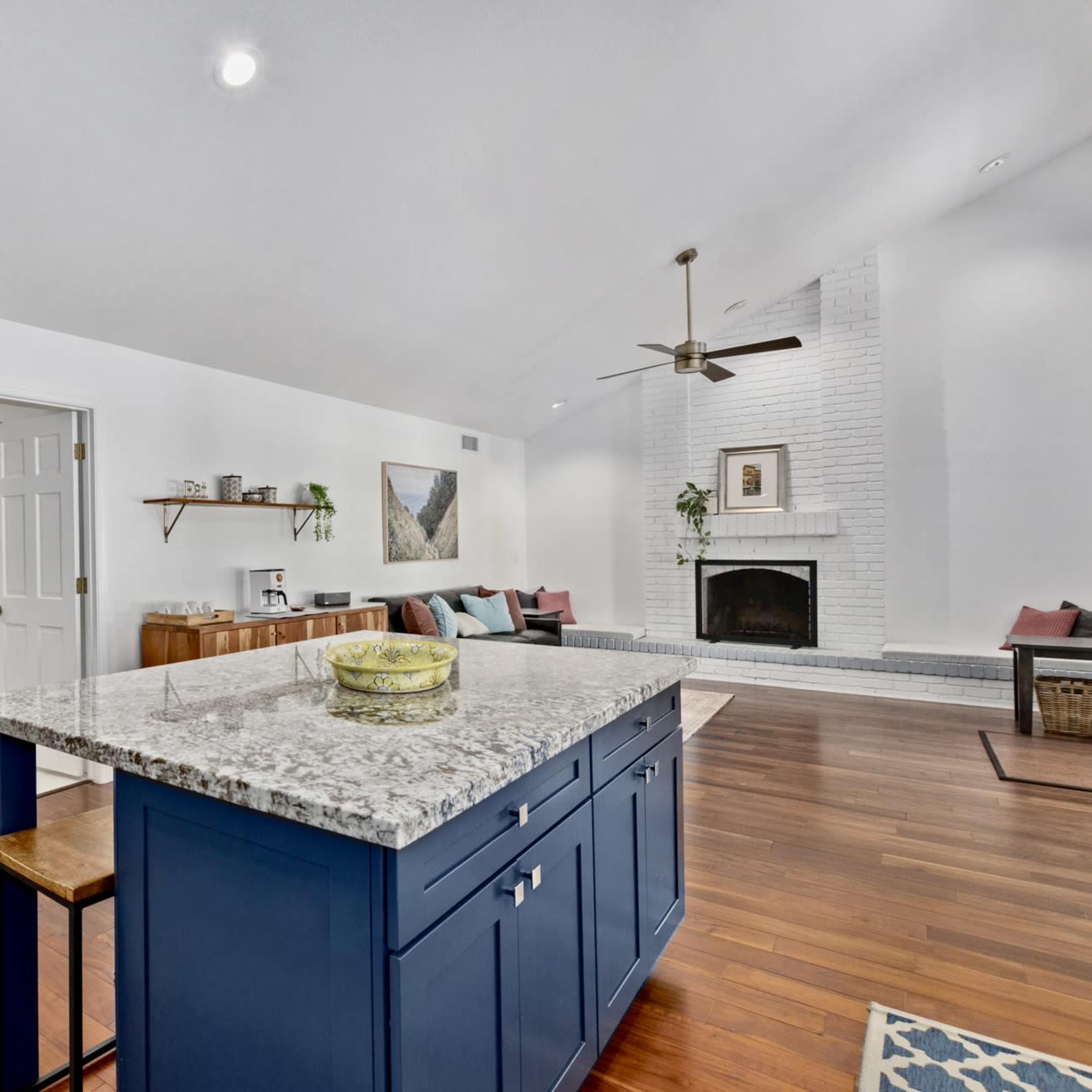 A modern kitchen features a blue island with granite countertops, adjacent to a cozy living area with a fireplace and seating.