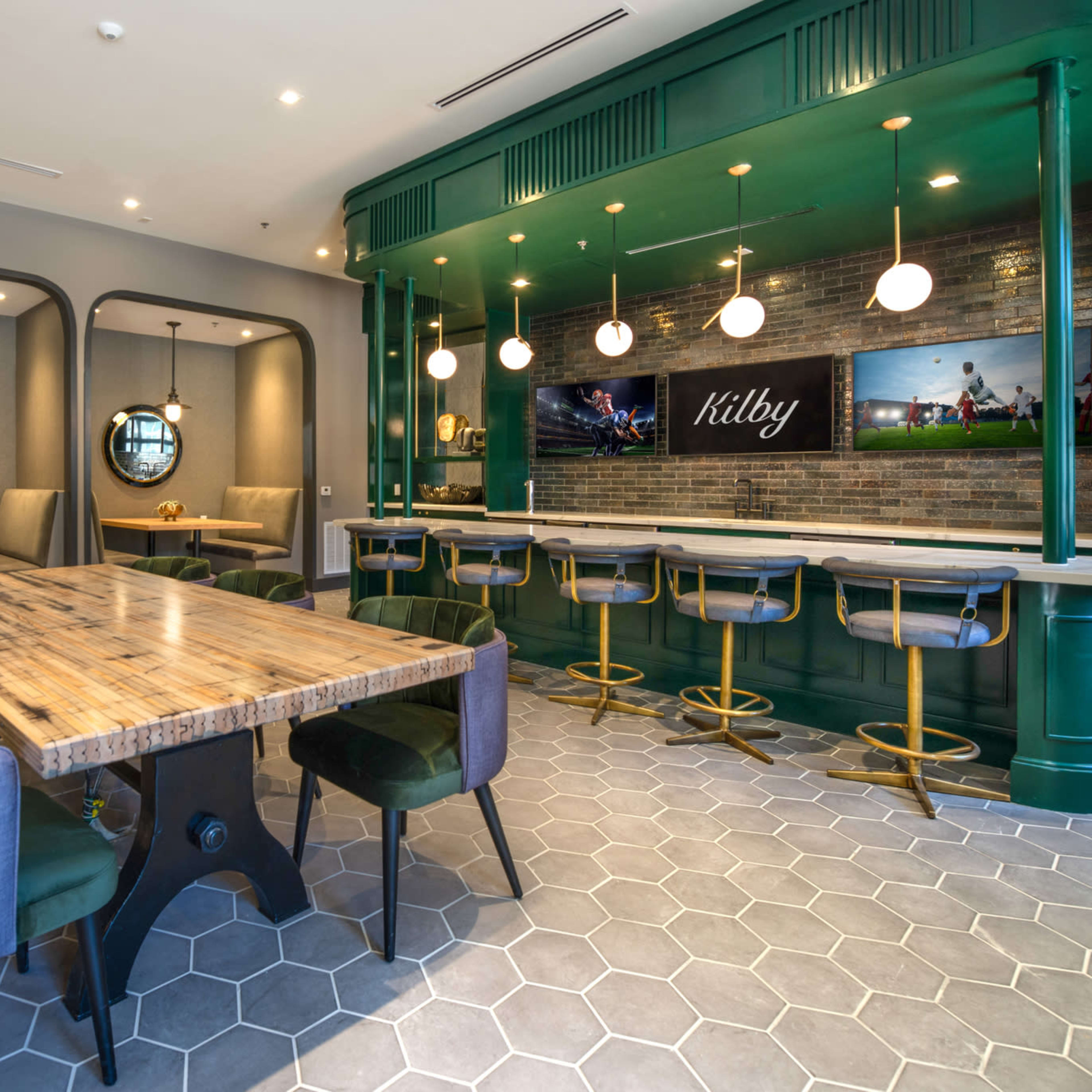 A modern dining area featuring a large wooden table surrounded by upholstered chairs and a bar with green accents and pendant lighting.
