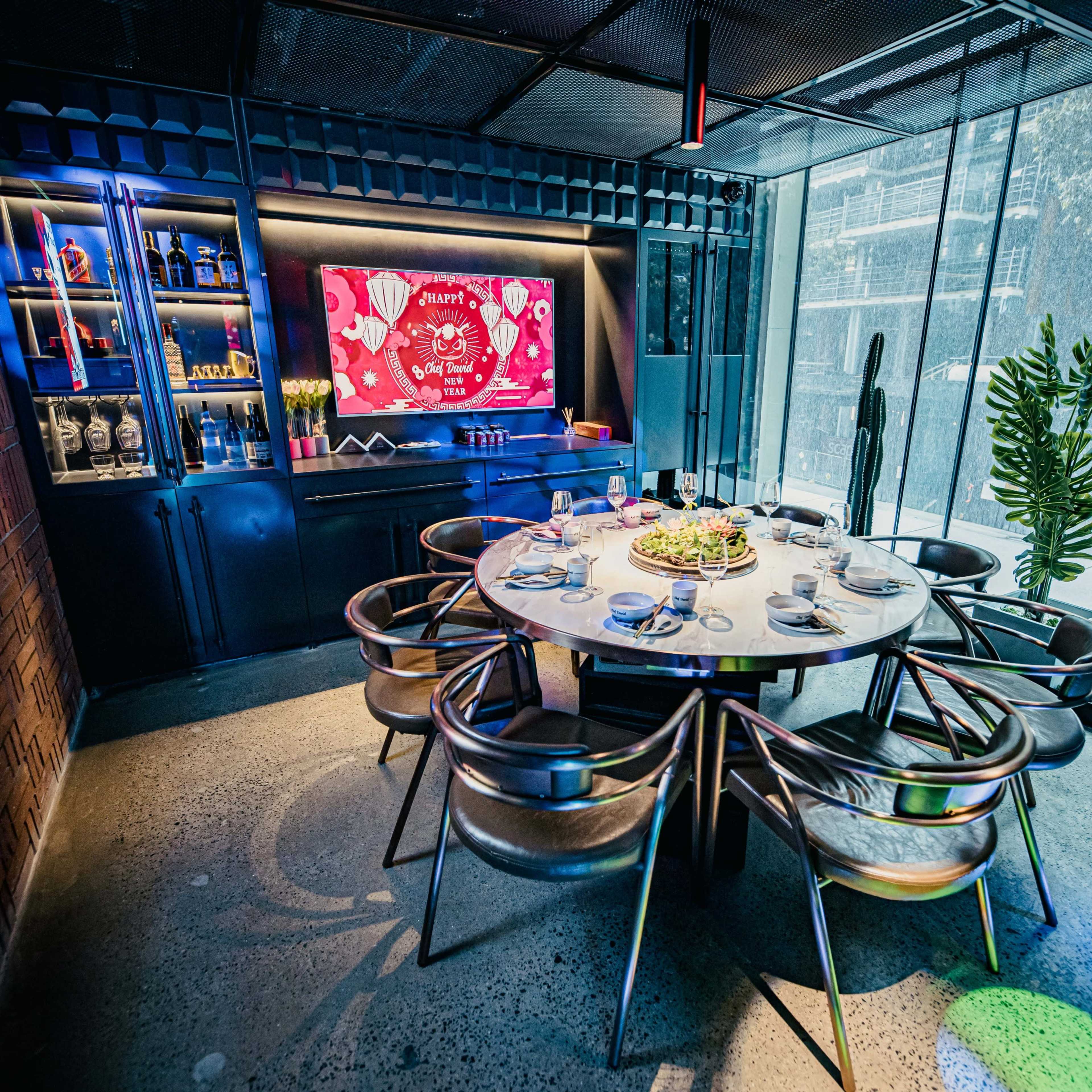 A round dining table is set with plates and cutlery in a modern restaurant featuring colorful lighting and a large screen on the wall.
