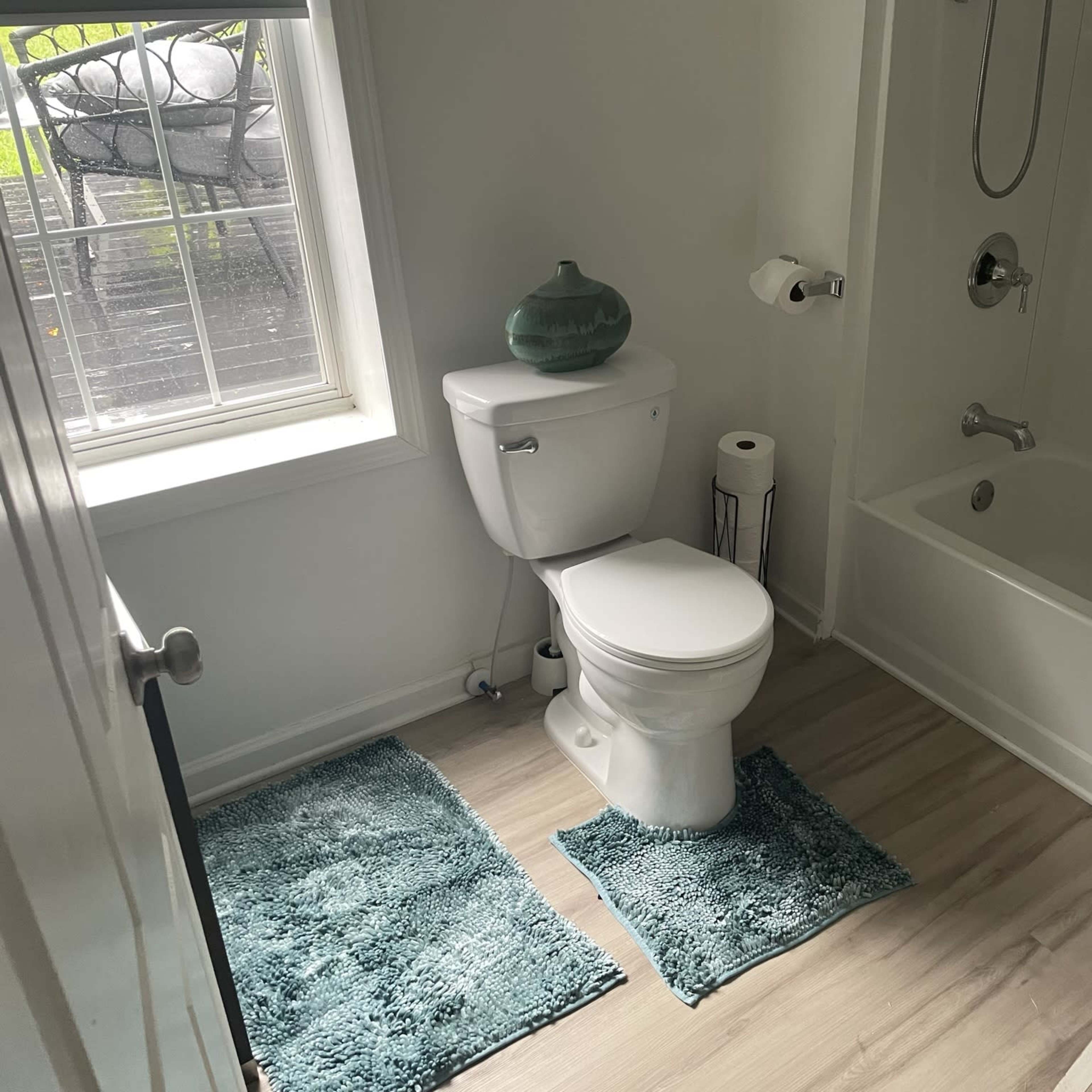 A bathroom featuring a toilet, a bathtub with a shower, and two teal bath mats on a light-colored wooden floor.