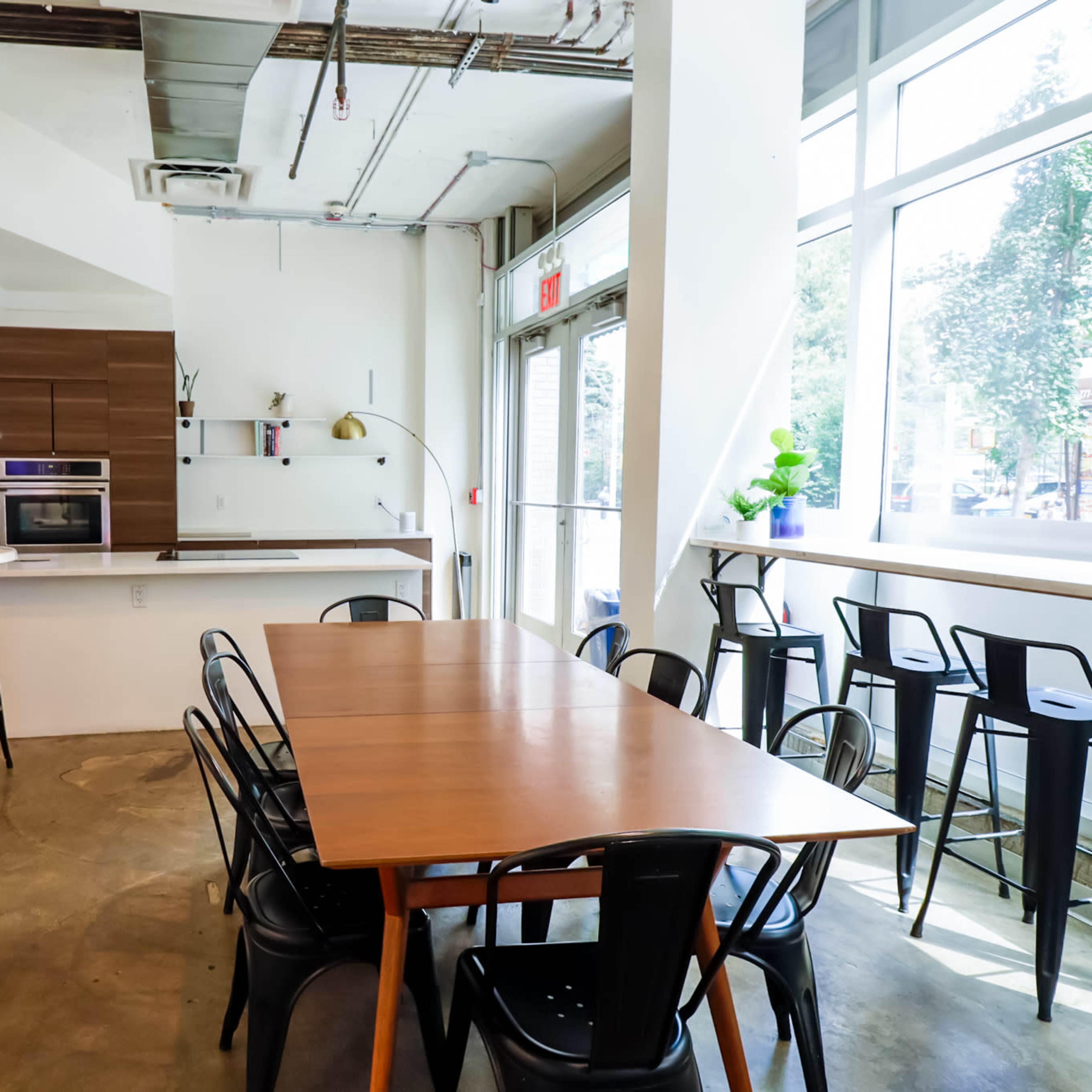 A modern, minimalist kitchen and dining area with a large wooden table surrounded by black chairs, set against a backdrop of large windows.