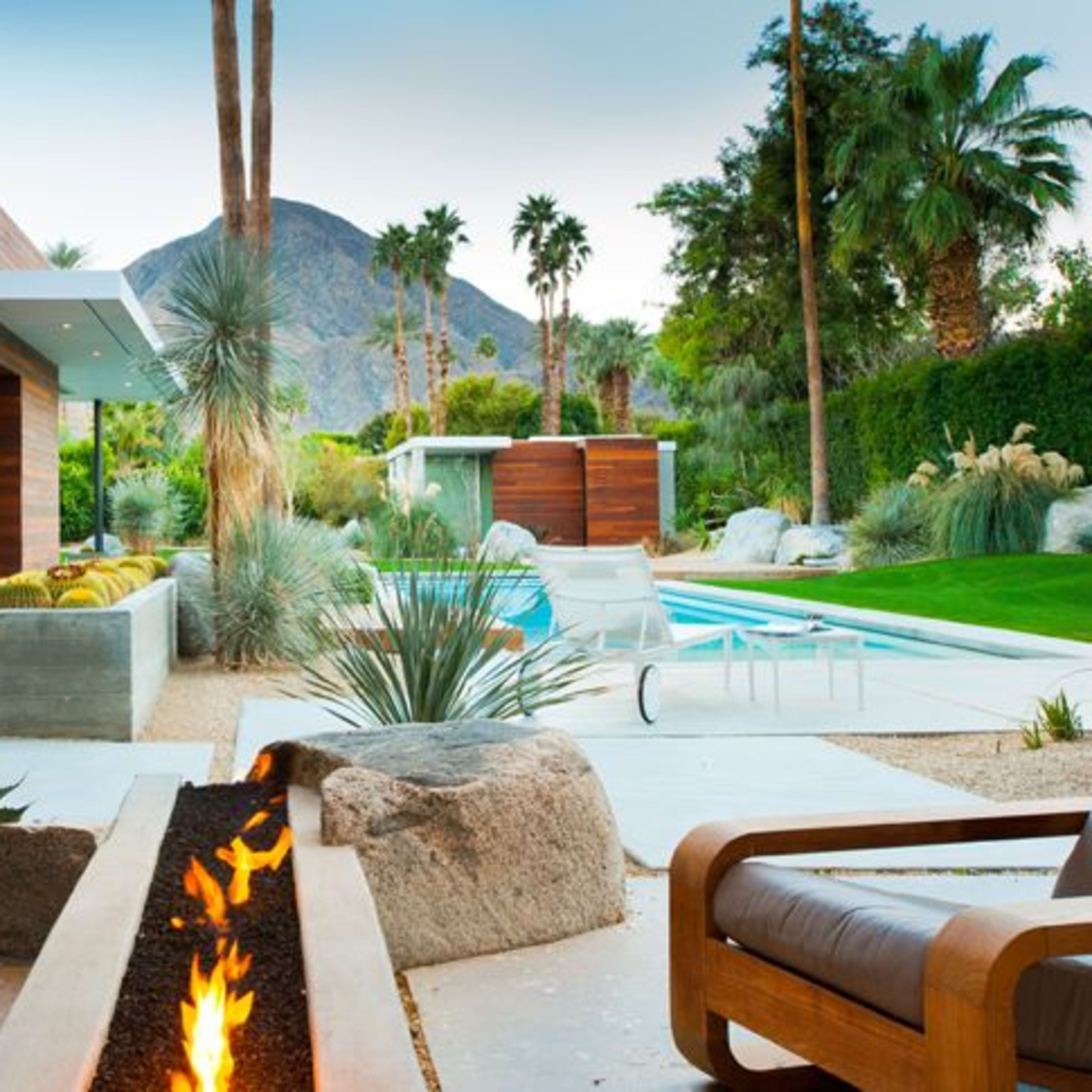 A modern patio features a fire pit surrounded by stylish chairs, with a swimming pool and palm trees in the background, set against a mountainous landscape.