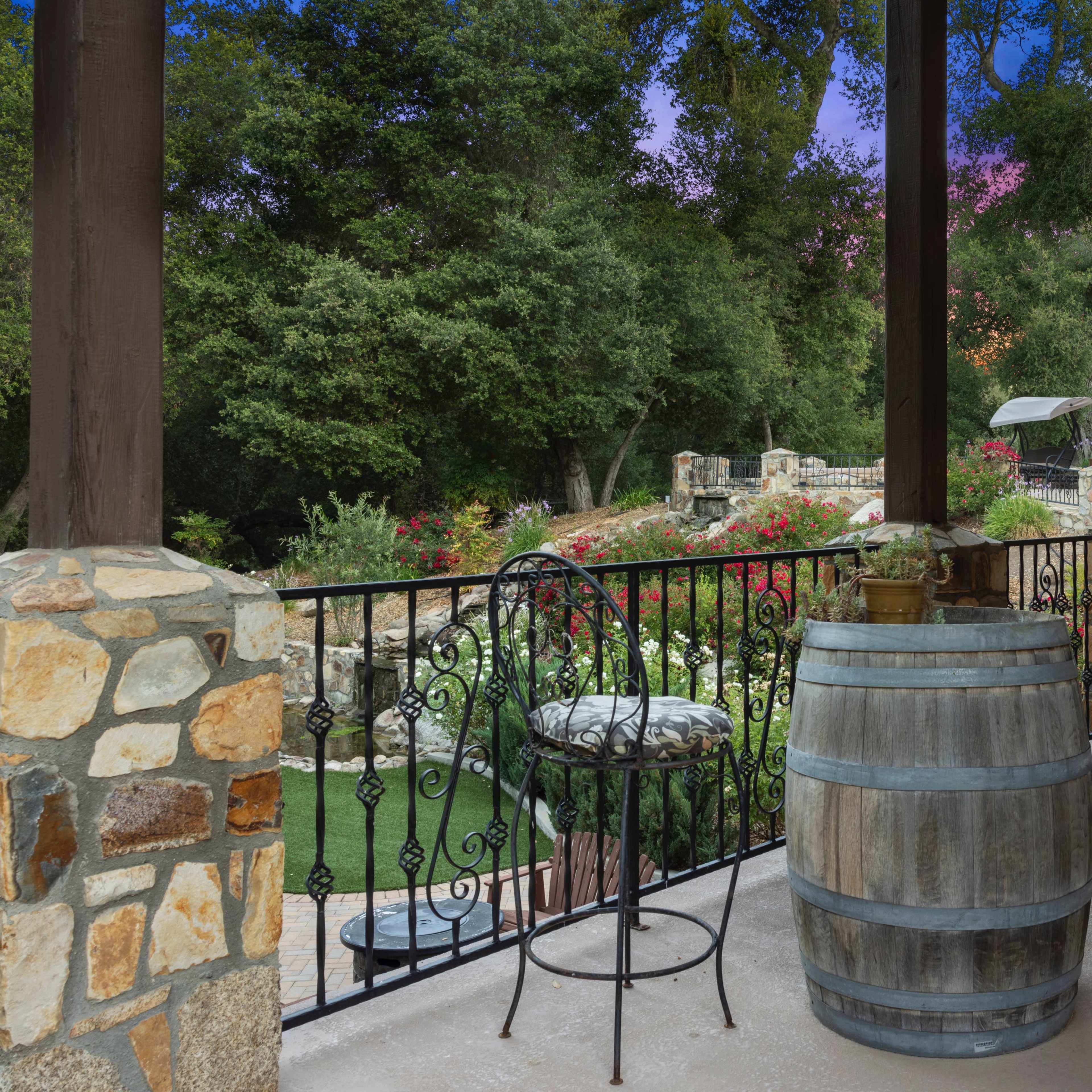 A covered outdoor patio with a stone railing, a wooden barrel table, and metal bar stools, overlooking a landscaped garden.