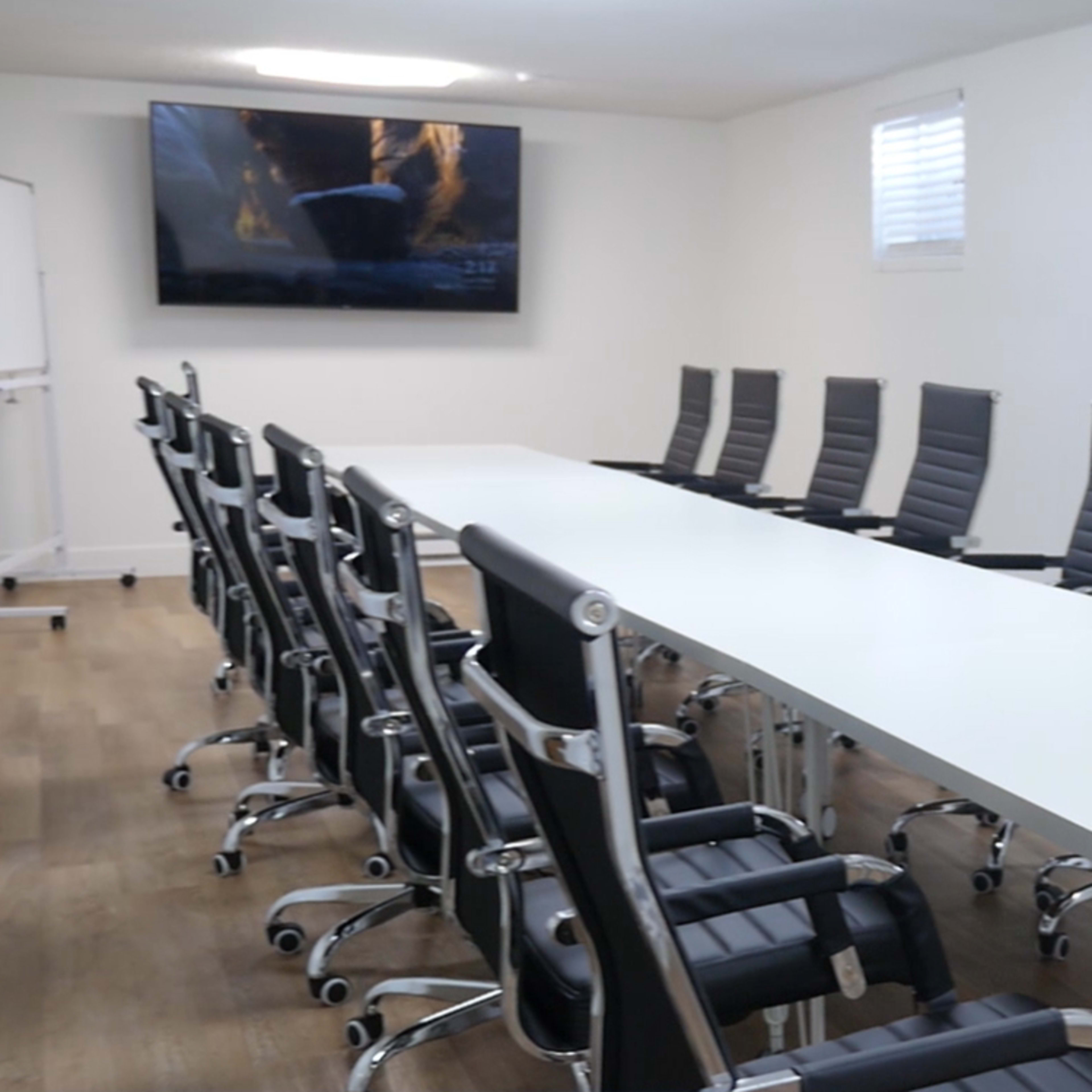 A long, white conference table is surrounded by twelve black ergonomic chairs in a well-lit meeting room with a wall-mounted screen.