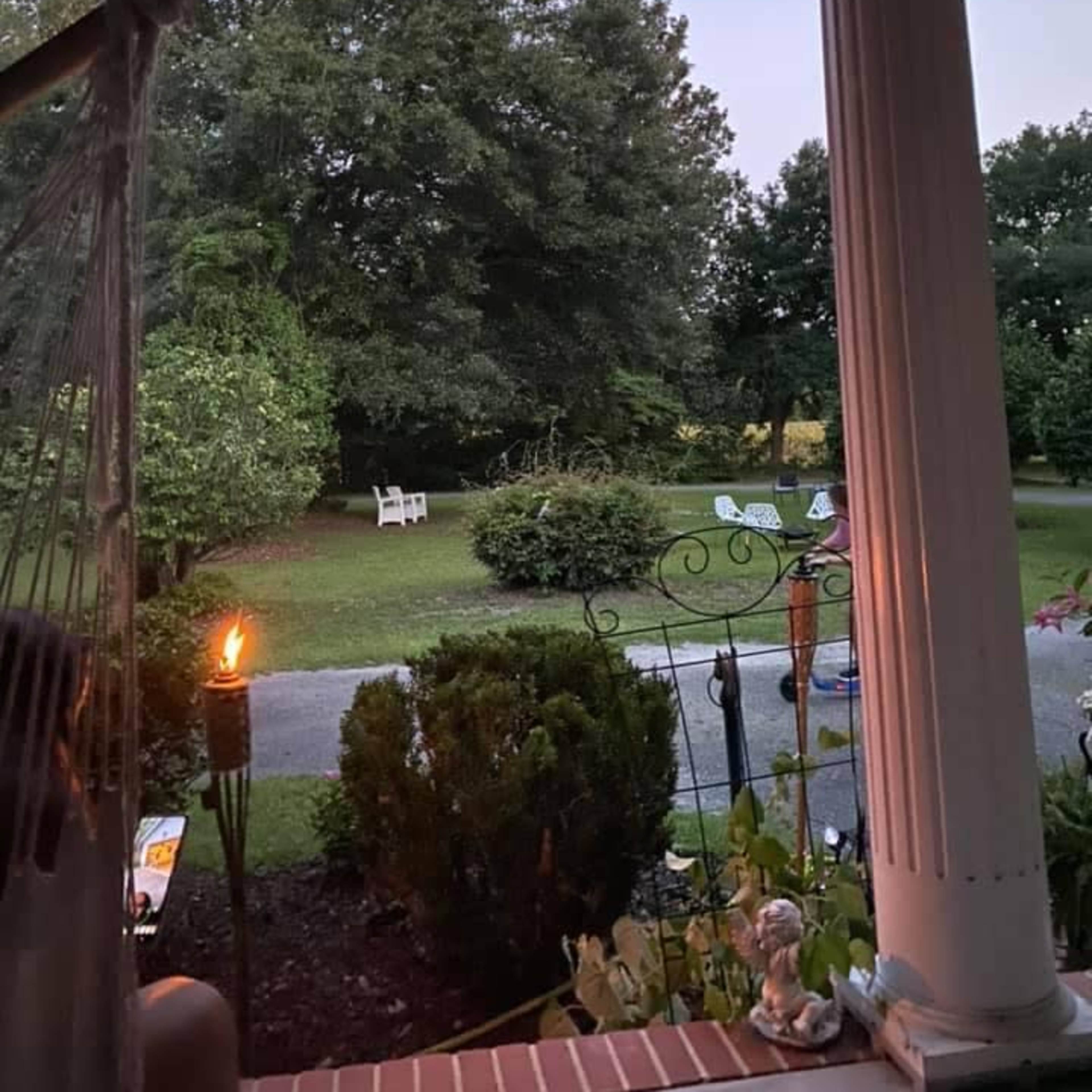 The view from a porch shows a garden with trees, a few chairs, and a lit torch in the foreground.