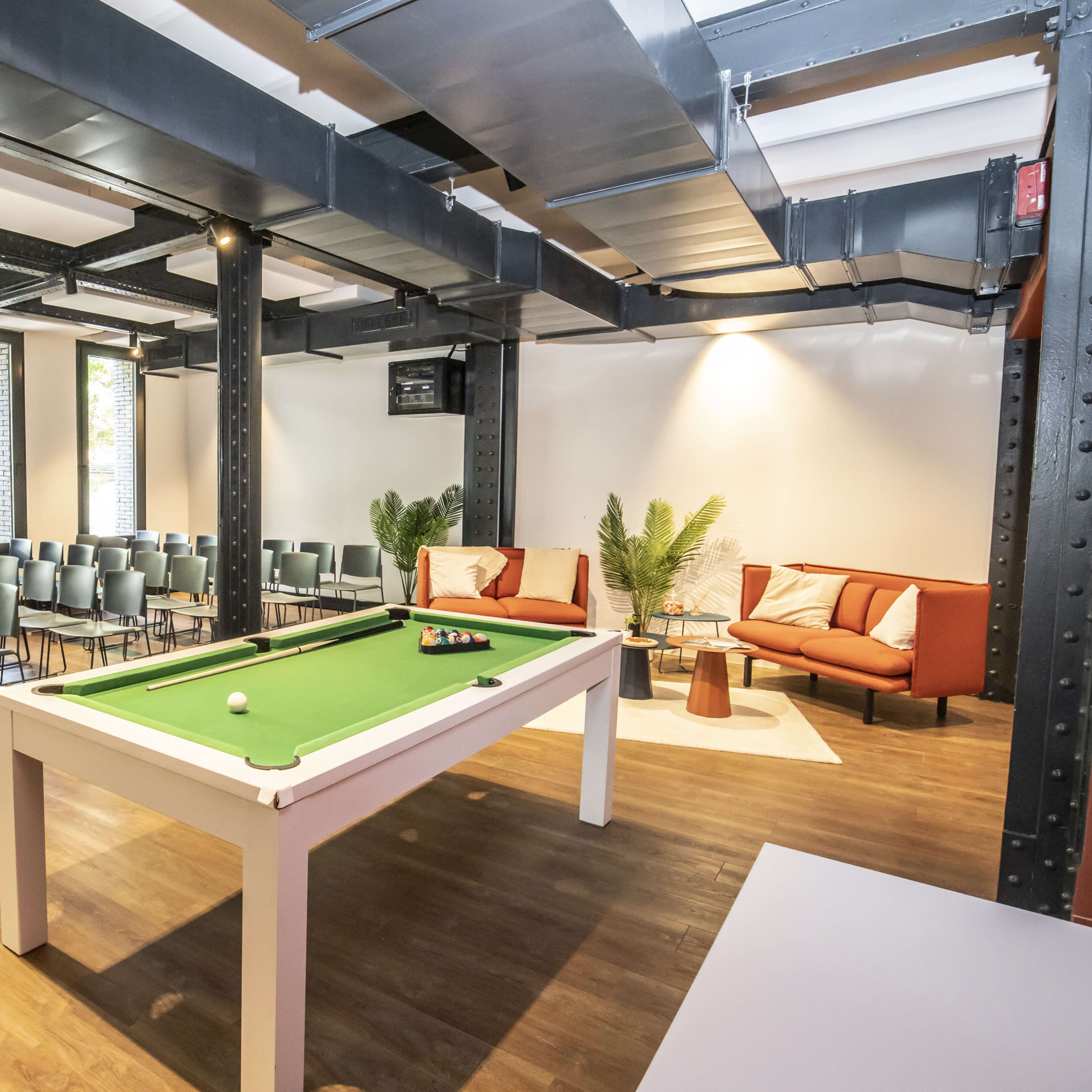 A spacious room features a green pool table centered among rows of metal chairs and a cozy seating area with two armchairs and a coffee table.