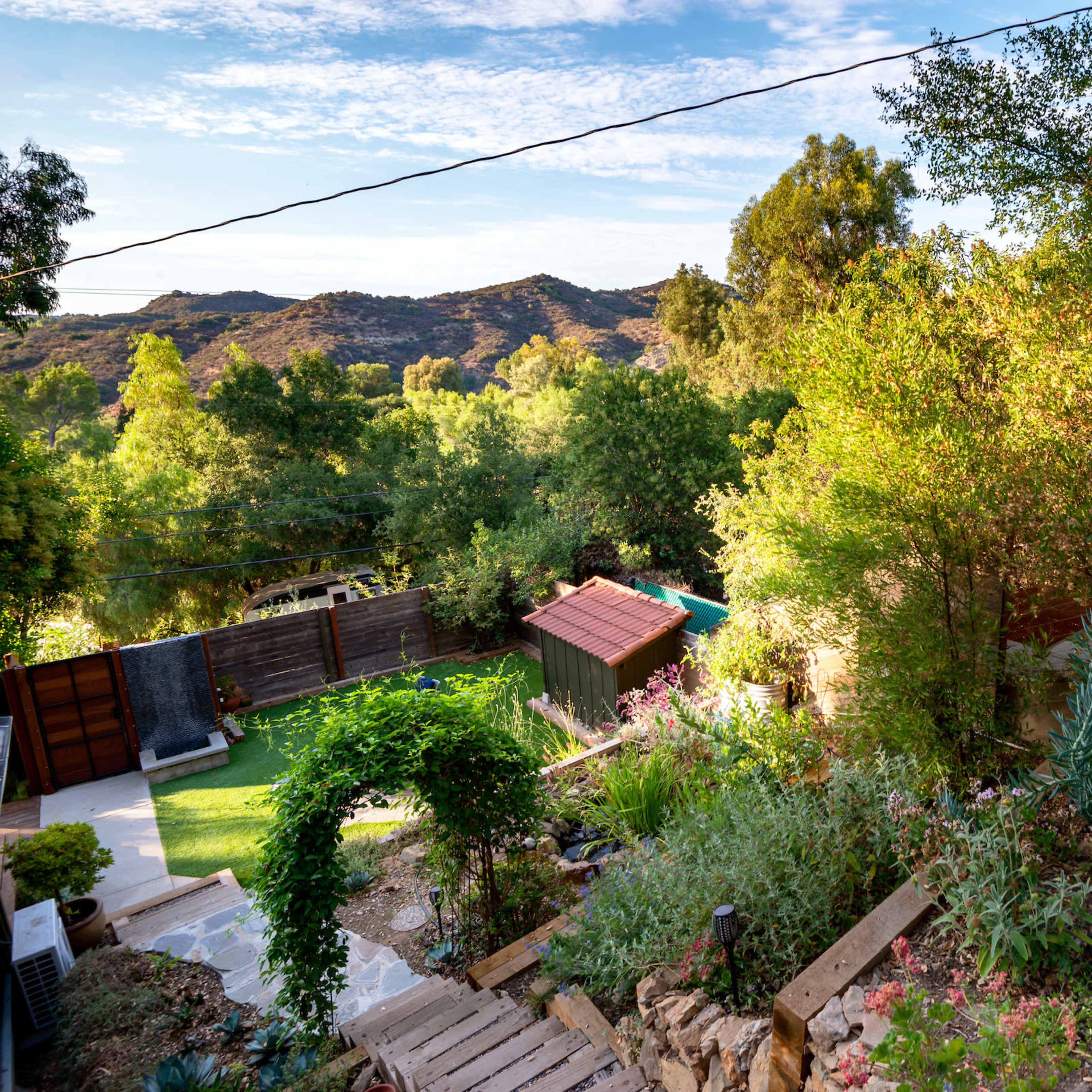 A well-maintained garden with various plants and a staircase leading down to a lush, green area, framed by hills in the background.