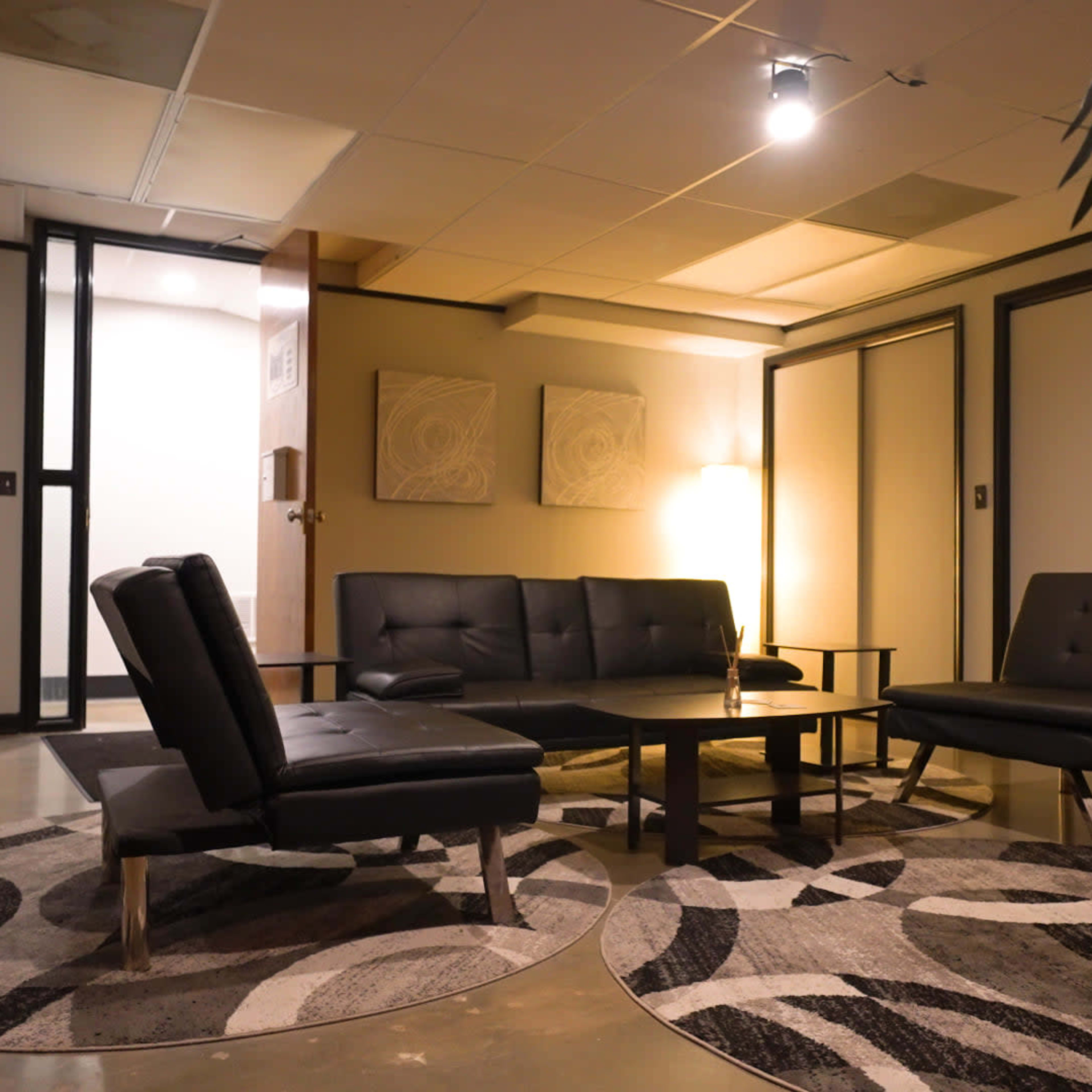A modern waiting area with black leather furniture, a round coffee table, and patterned area rugs, illuminated by overhead lighting.