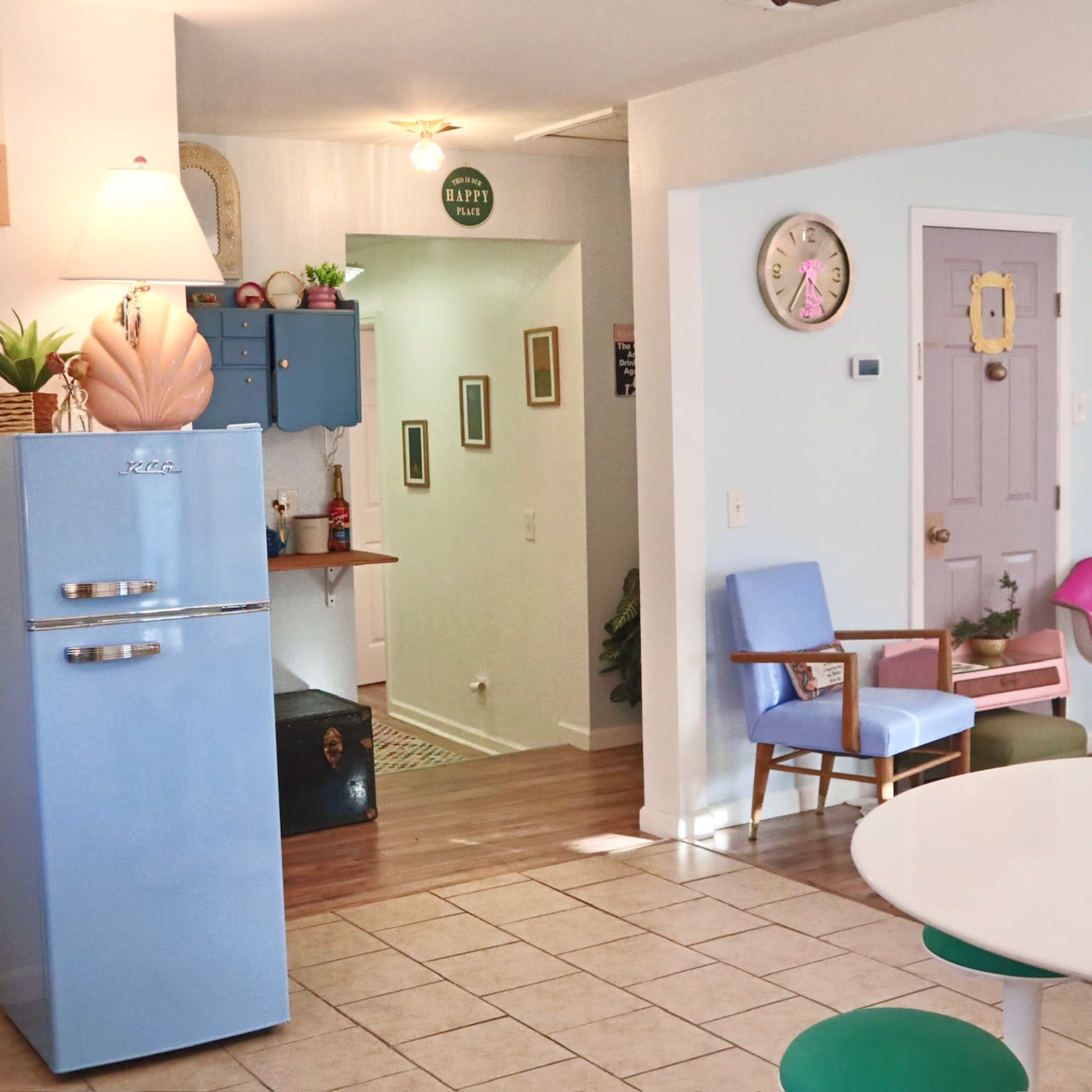 A brightly lit indoor space featuring a blue vintage refrigerator, a white round dining table, and colorful chairs alongside light-colored walls.