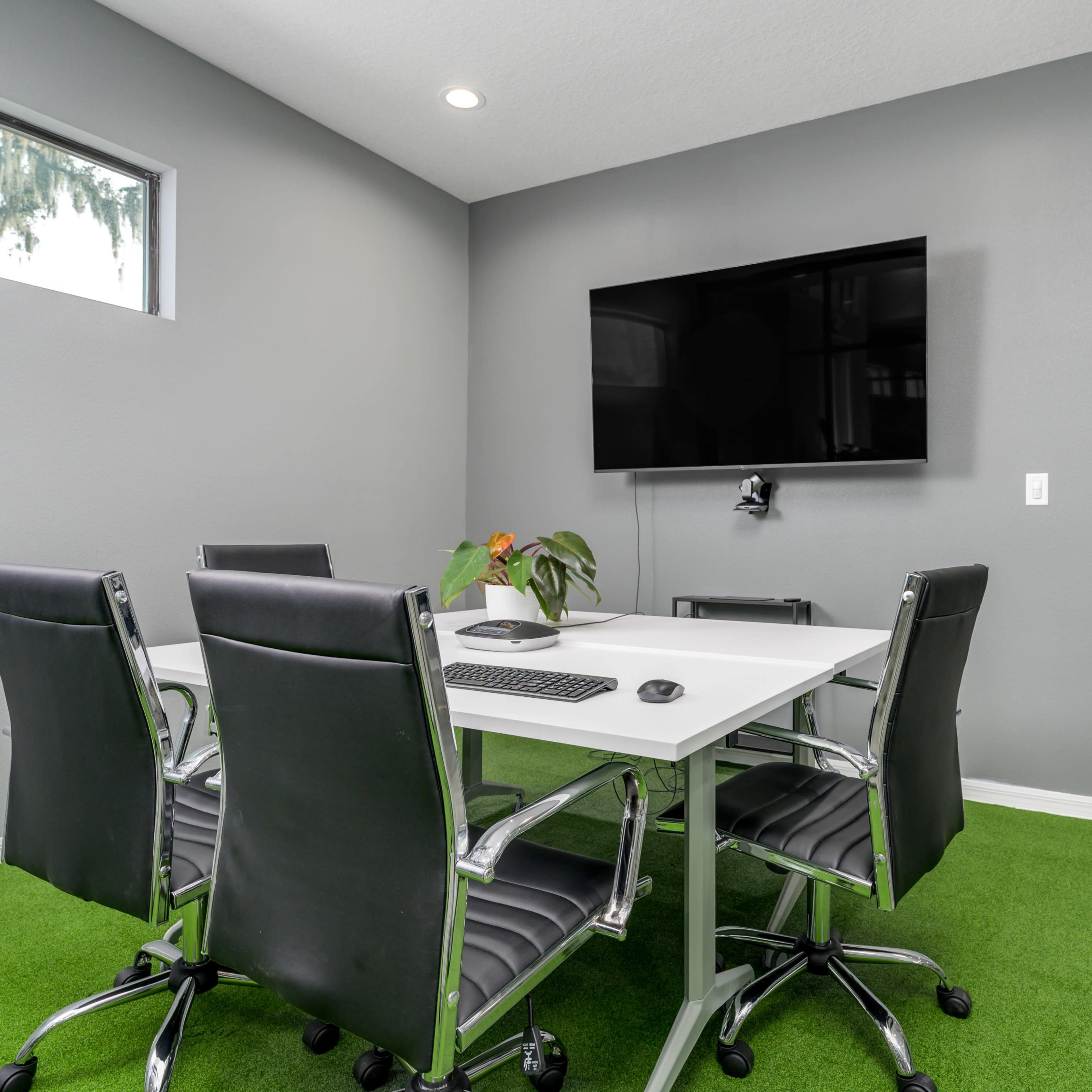 A conference room with a white table, black chairs, a green carpet, and a mounted television on a gray wall.