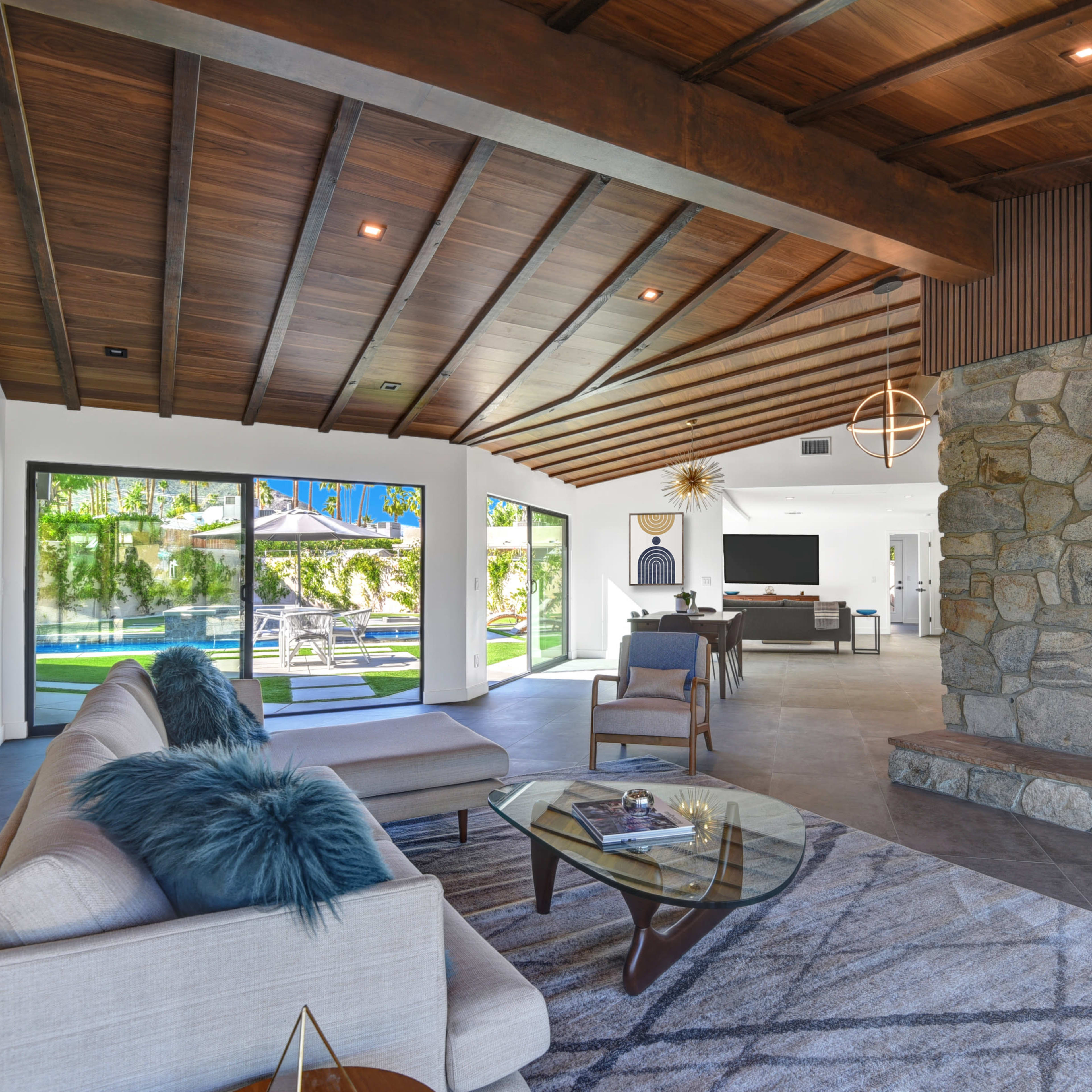 A modern living room features a stone fireplace, wooden ceiling beams, and large glass doors opening to an outdoor space.