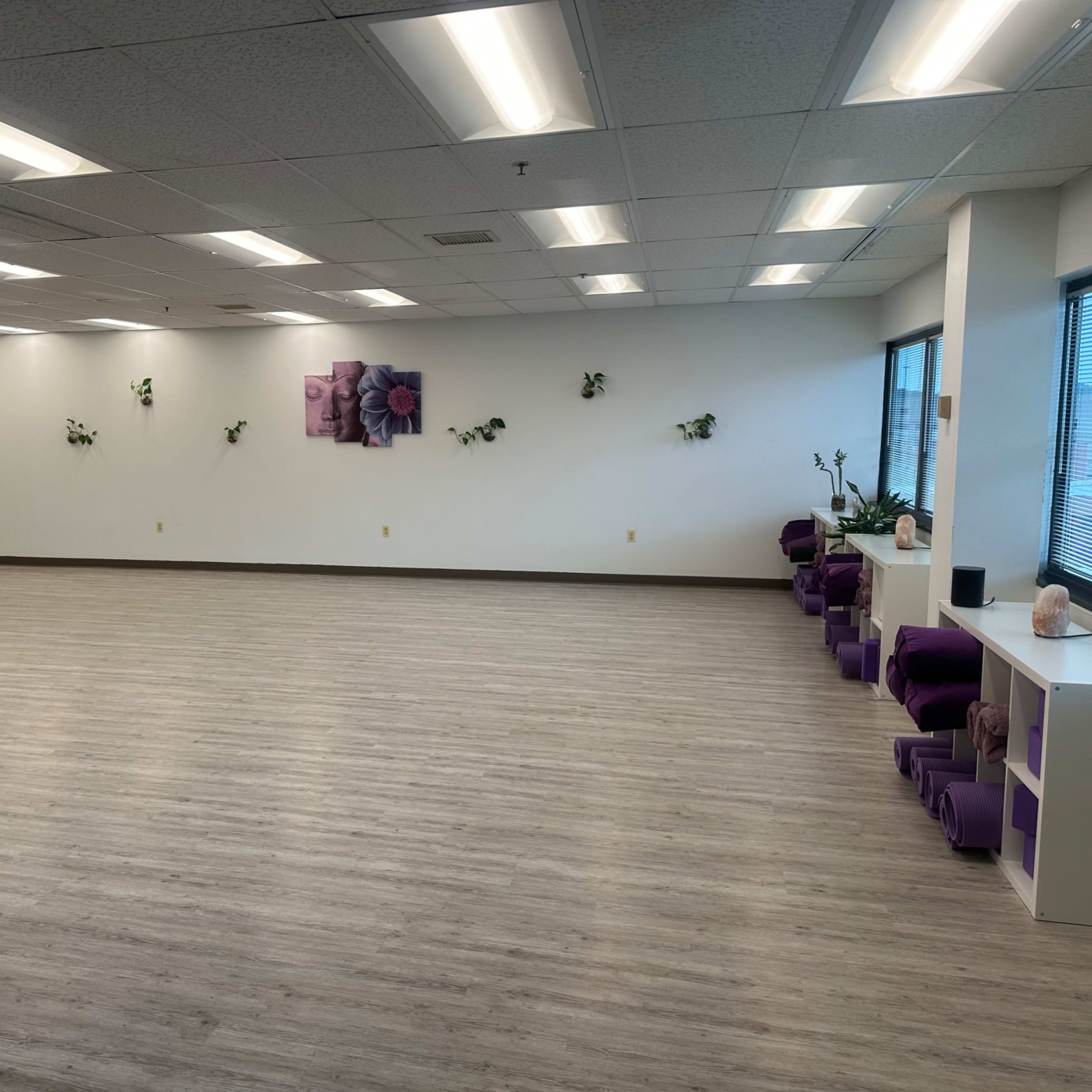 A spacious, empty room with light wood flooring, purple seating against the wall, and plants along the edges.