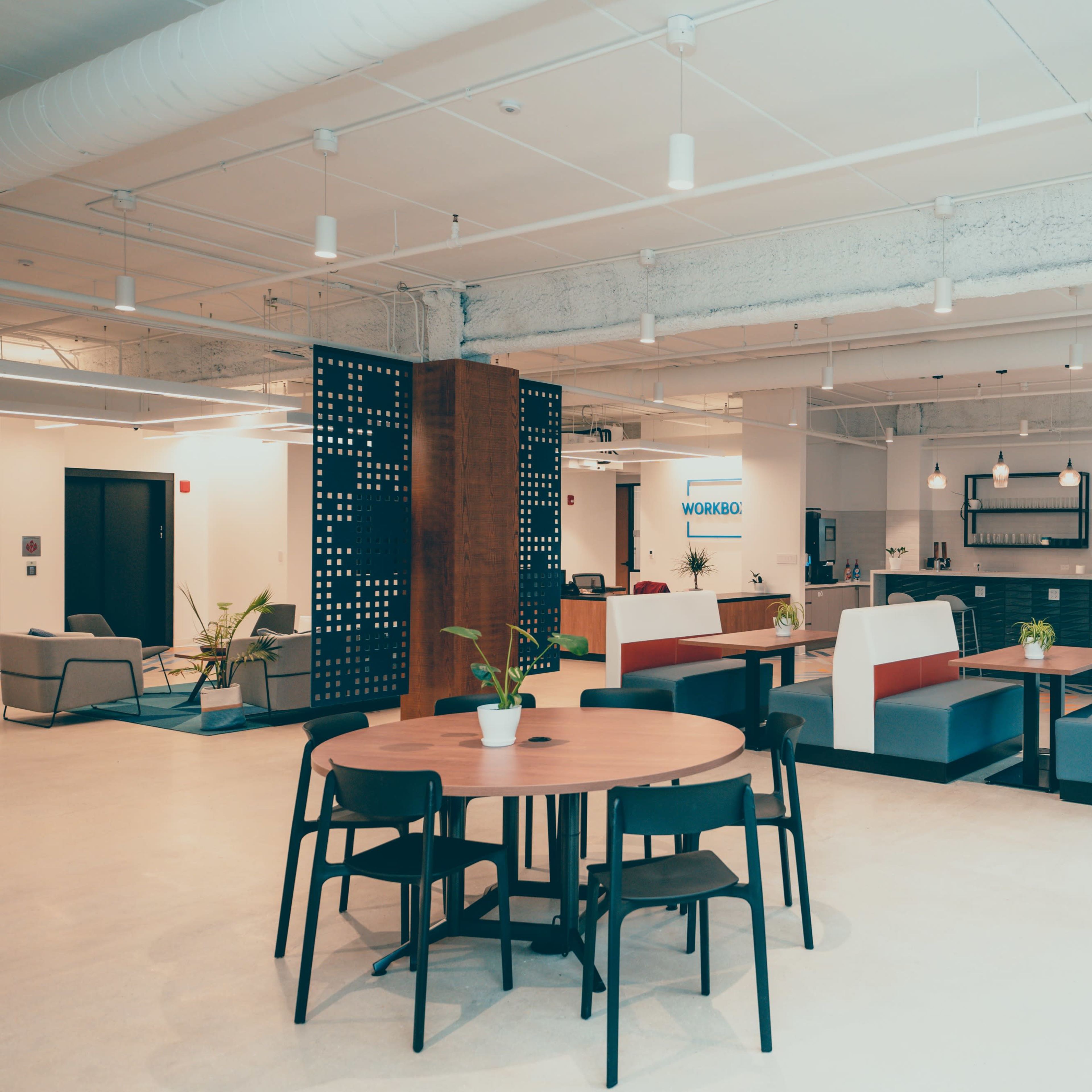 A modern office space featuring a kitchen area, seating booths, and a dining table surrounded by black chairs.