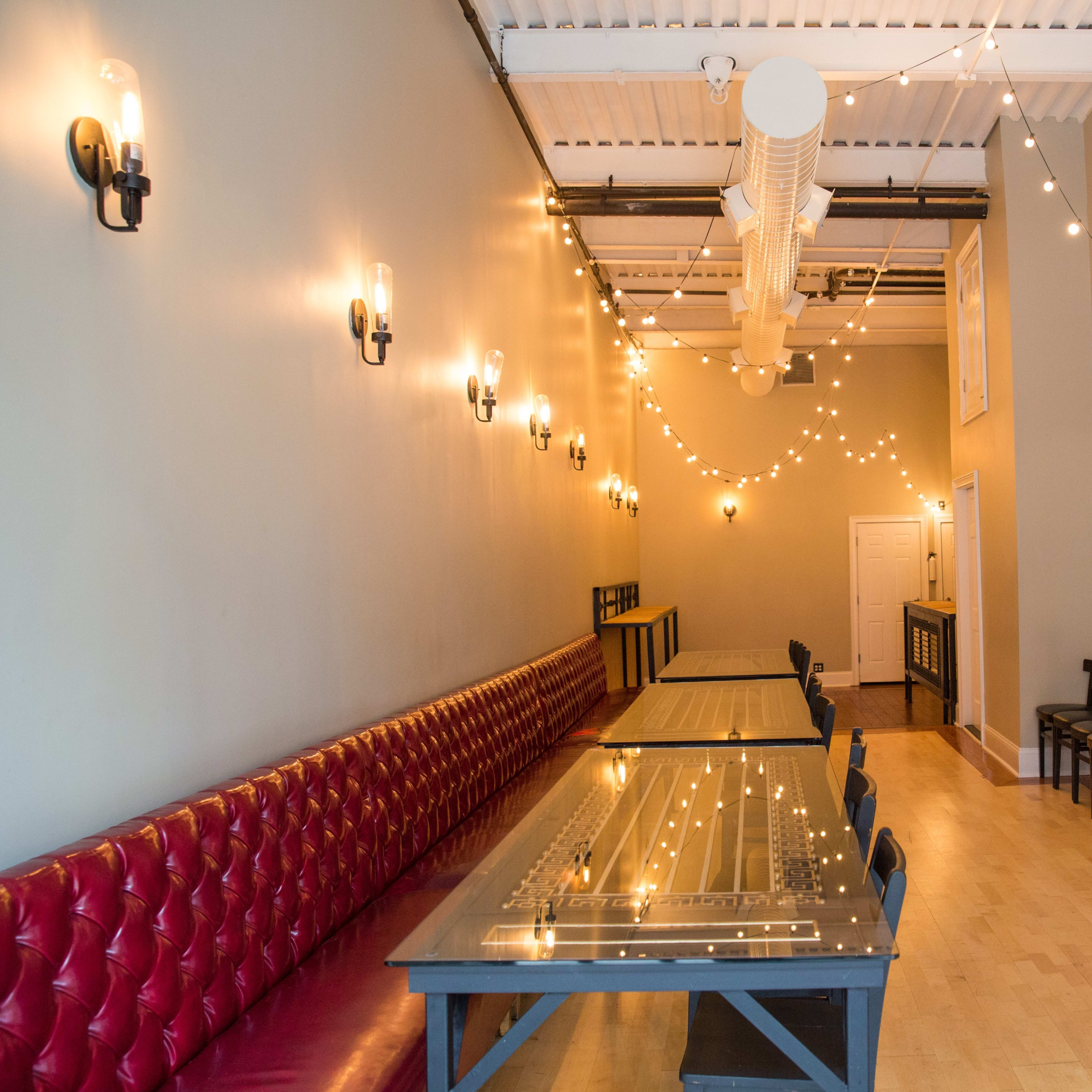 A long, narrow room with red tufted seating on one side, wooden tables in the center, and string lights hanging from the ceiling.