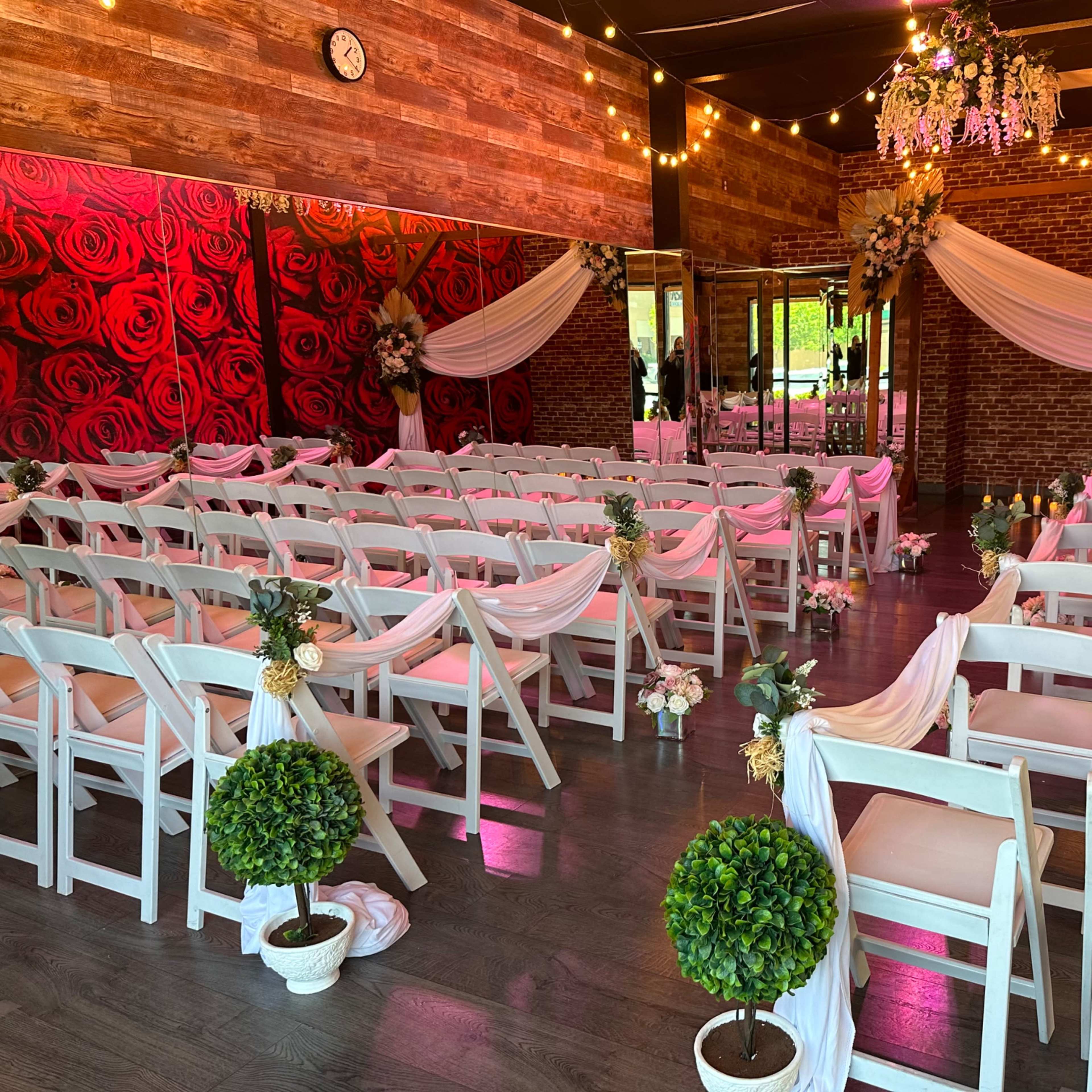 A decorated event space features rows of white chairs with floral accents, arranged facing a small altar, surrounded by rose-themed decor and soft lighting.