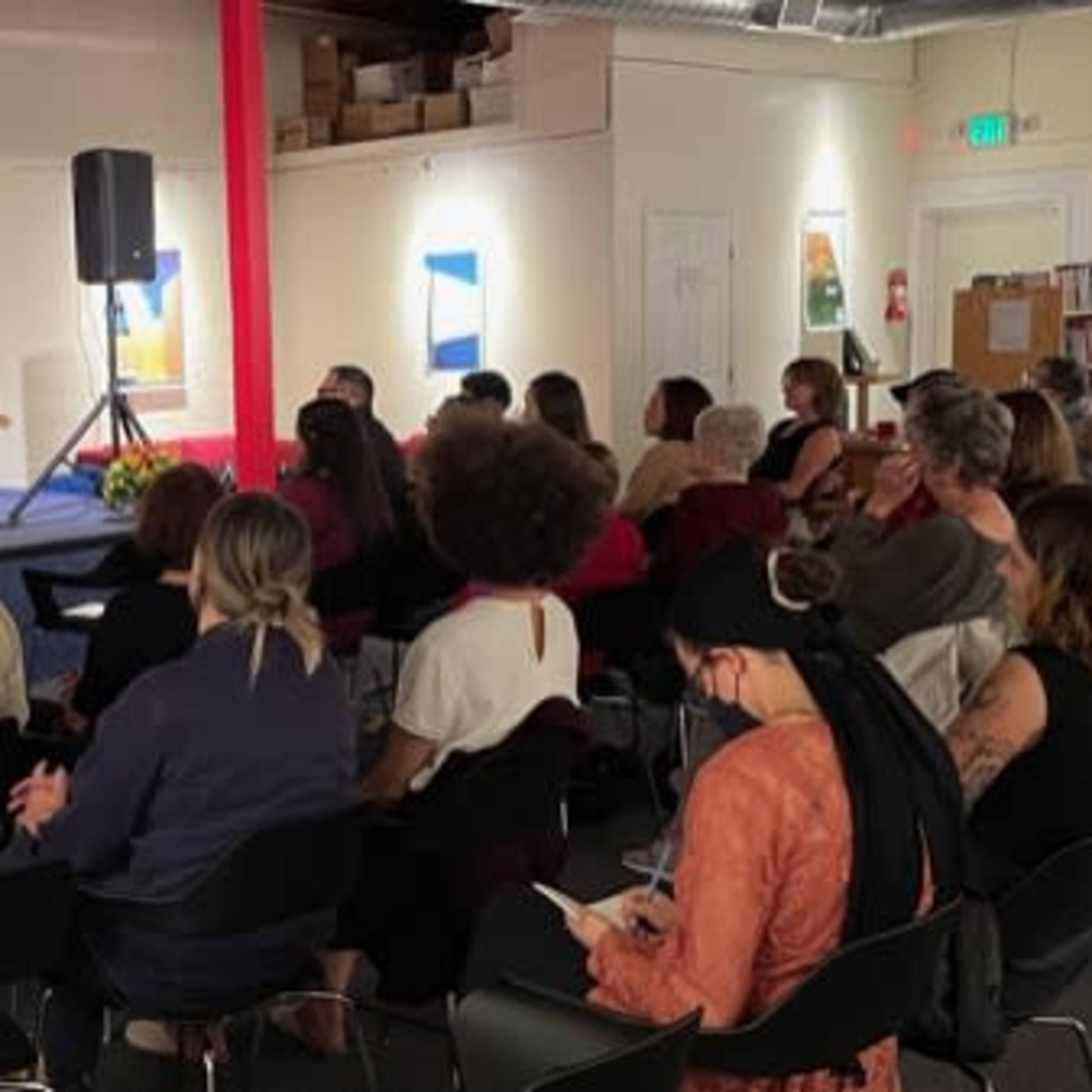 A speaker sits on a stage speaking to an audience in a seated arrangement at a public event in a gallery.