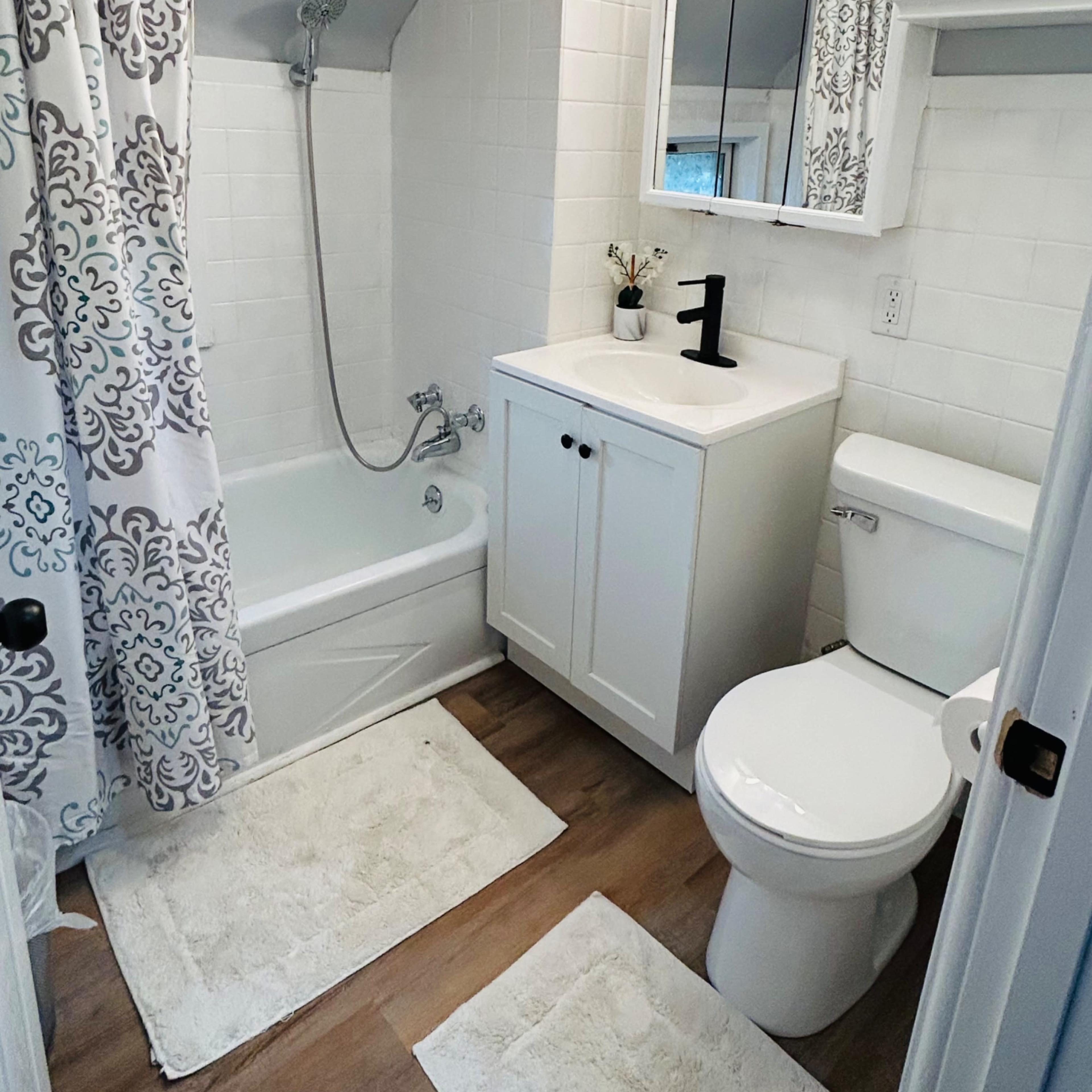A compact bathroom featuring a bathtub with a shower, a white vanity with a sink, a toilet, and decorative elements on the walls.