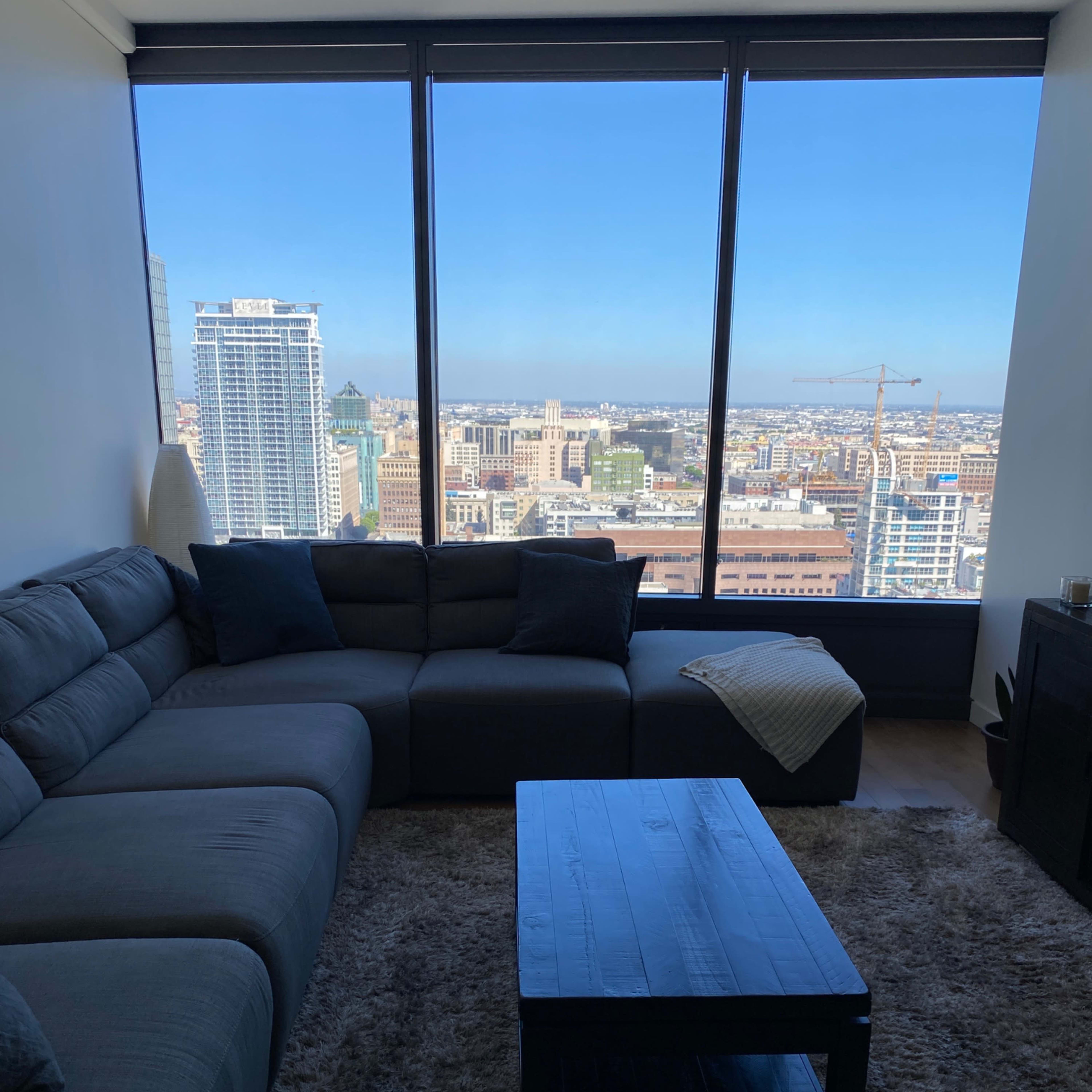 A modern living room features a gray sectional sofa and a black coffee table with a city skyline visible through large floor-to-ceiling windows.