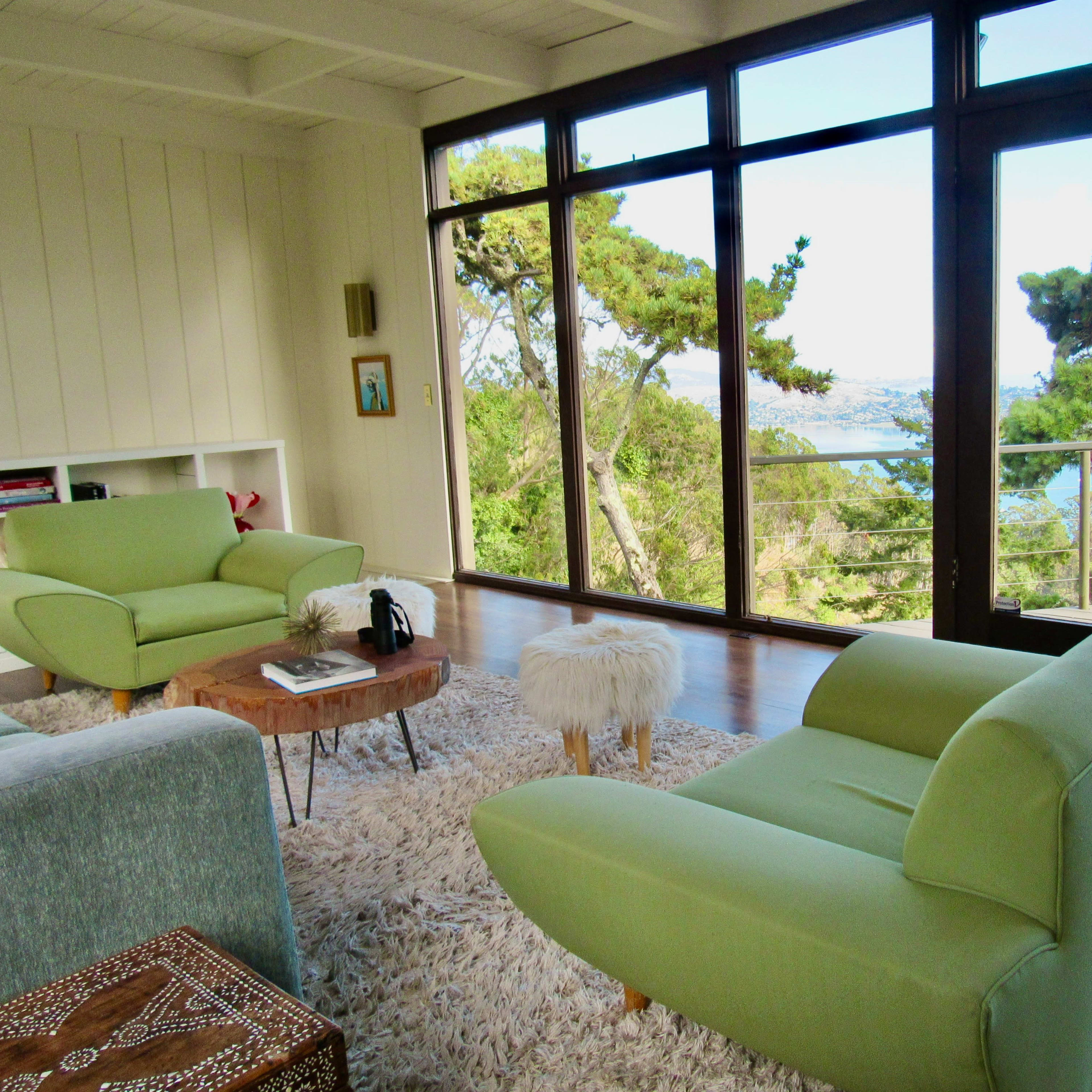 A brightly lit room with two green chairs, a coffee table, and large windows offering a view of trees and water.