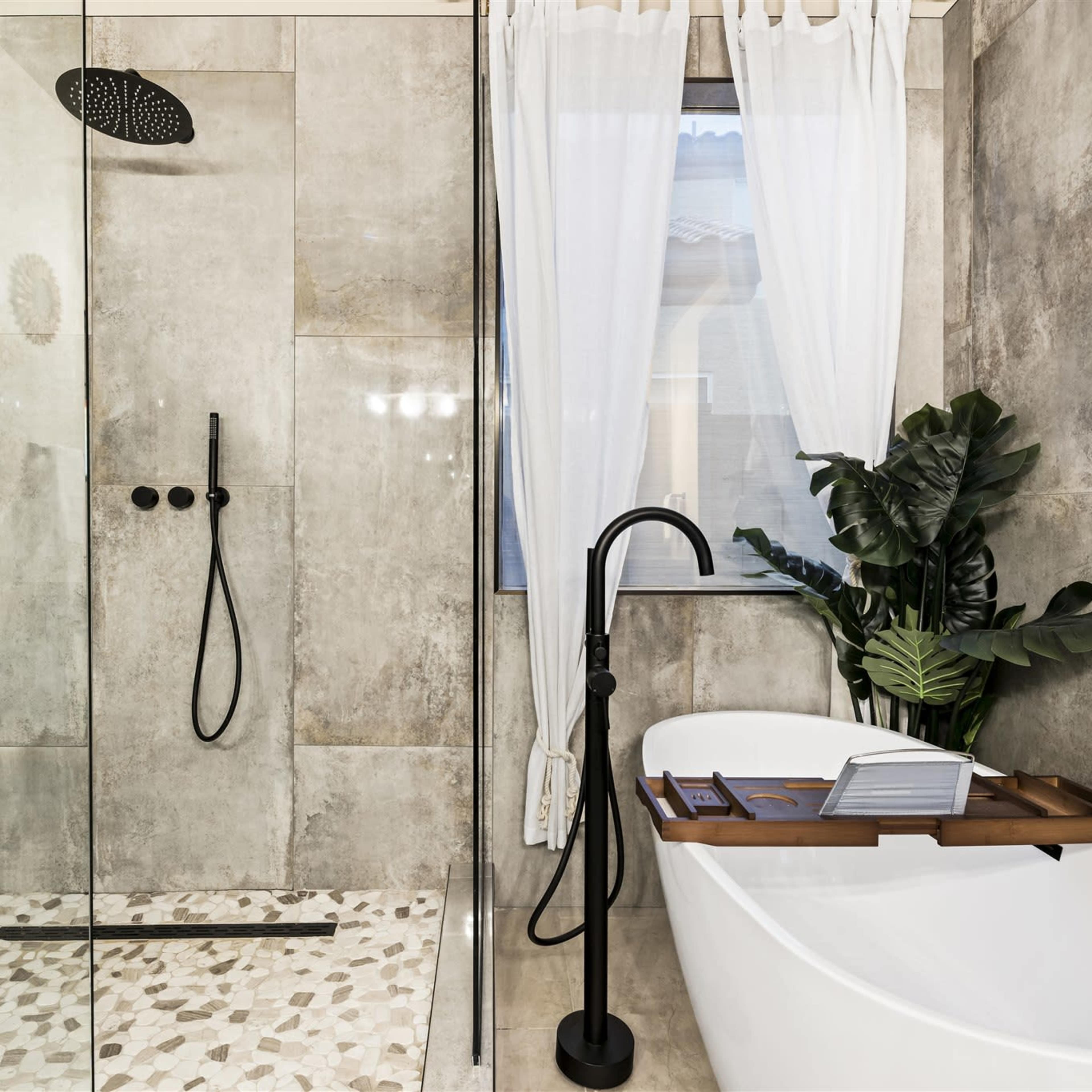 A modern bathroom featuring a glass shower, a freestanding bathtub, and a potted plant near a window with sheer curtains.