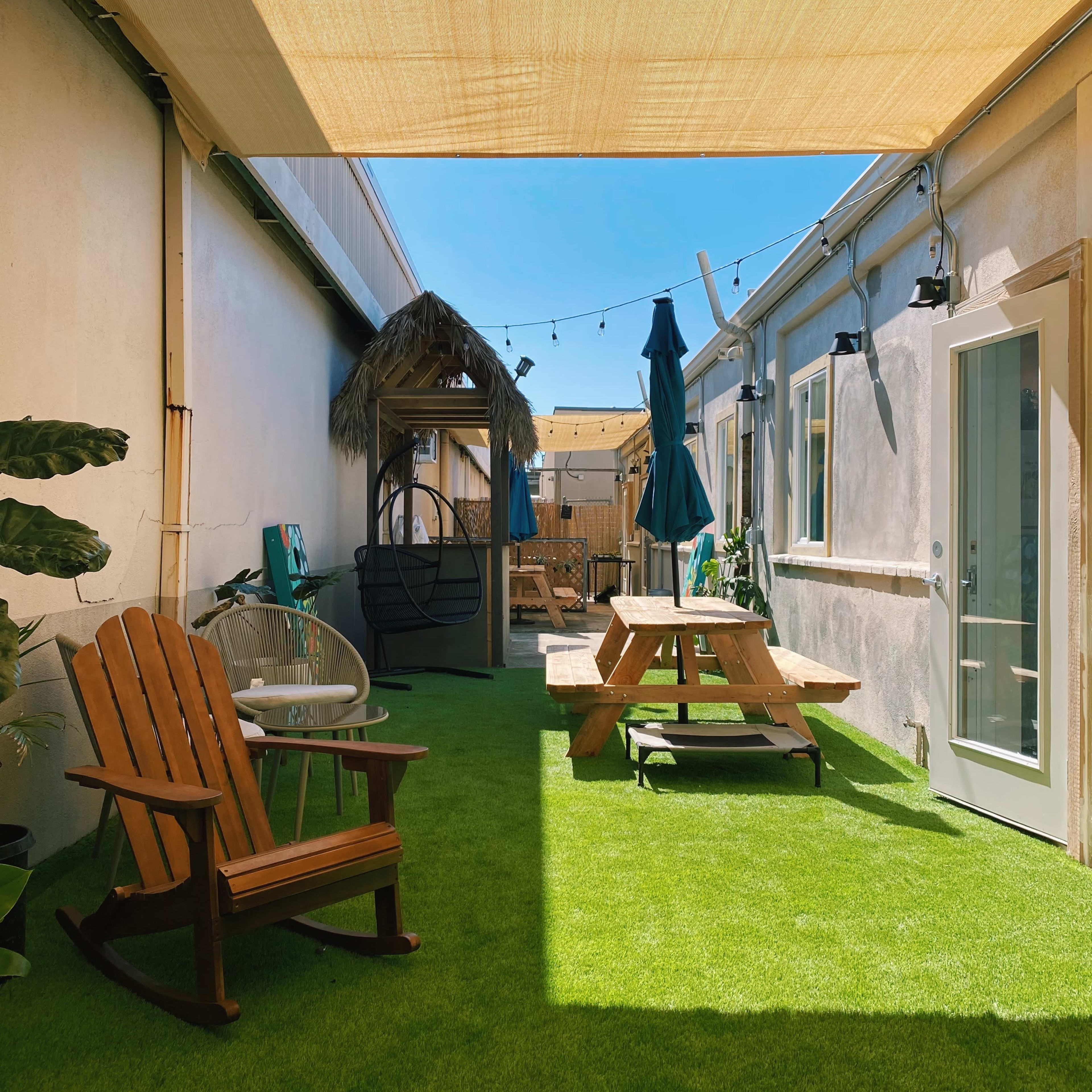 A well-lit outdoor courtyard with artificial grass, featuring a rocking chair, a picnic table, a shaded area with a thatched roof, and string lights.