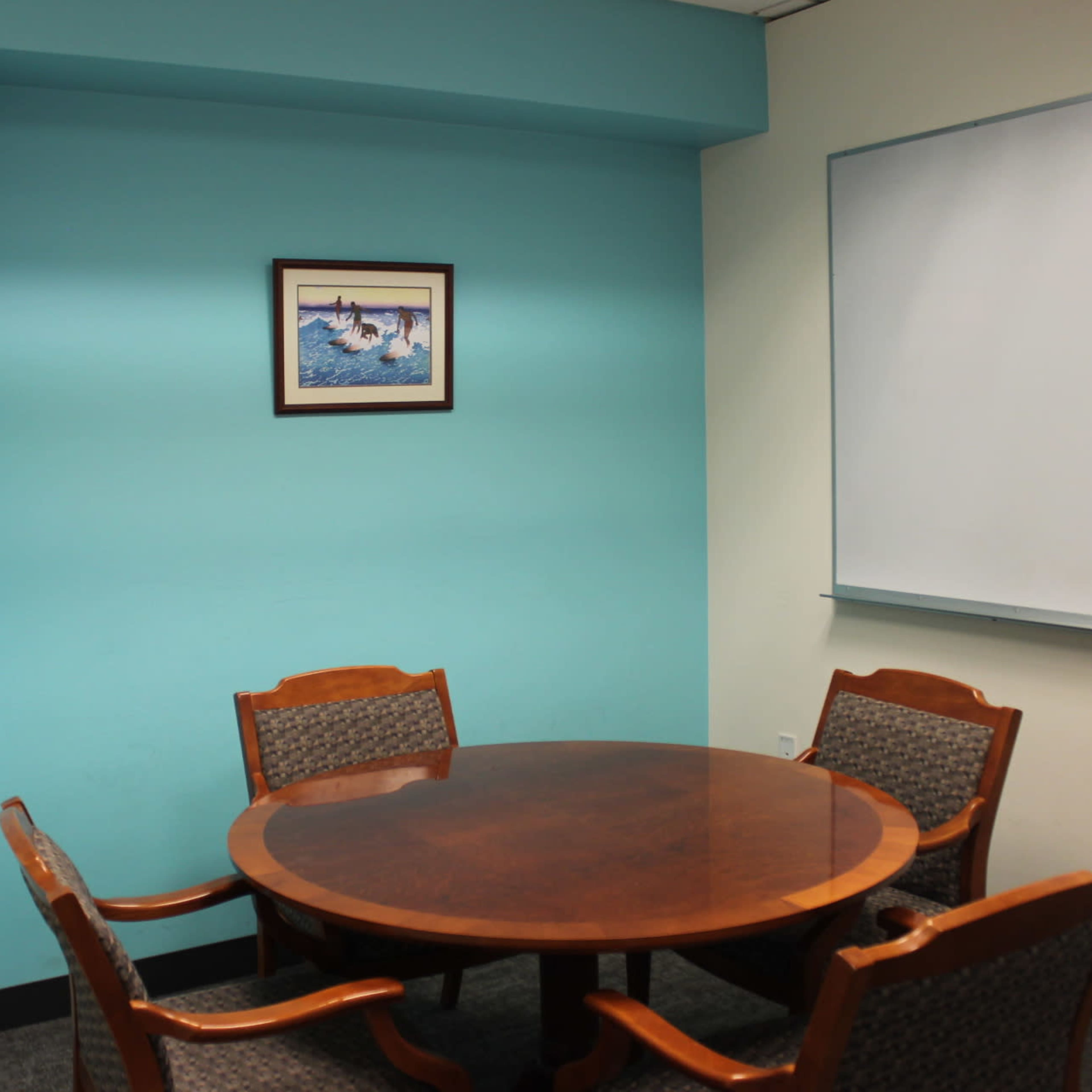 A round wooden table surrounded by four wooden chairs is positioned in a turquoise-colored meeting room with a framed picture on the wall and a whiteboard.