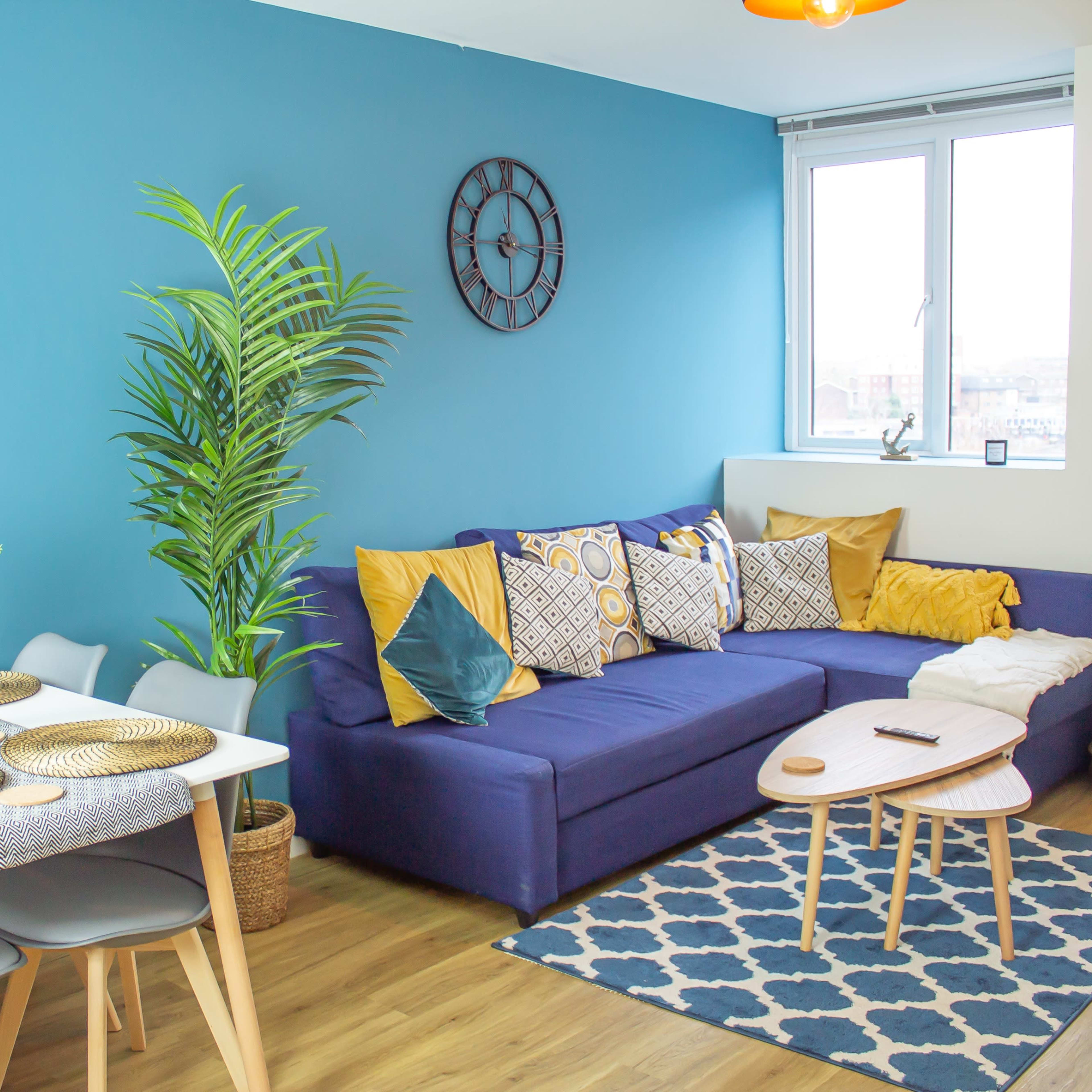 A bright living room features a blue wall, a couch with decorative pillows, a dining table set for four, and a large window allowing natural light.