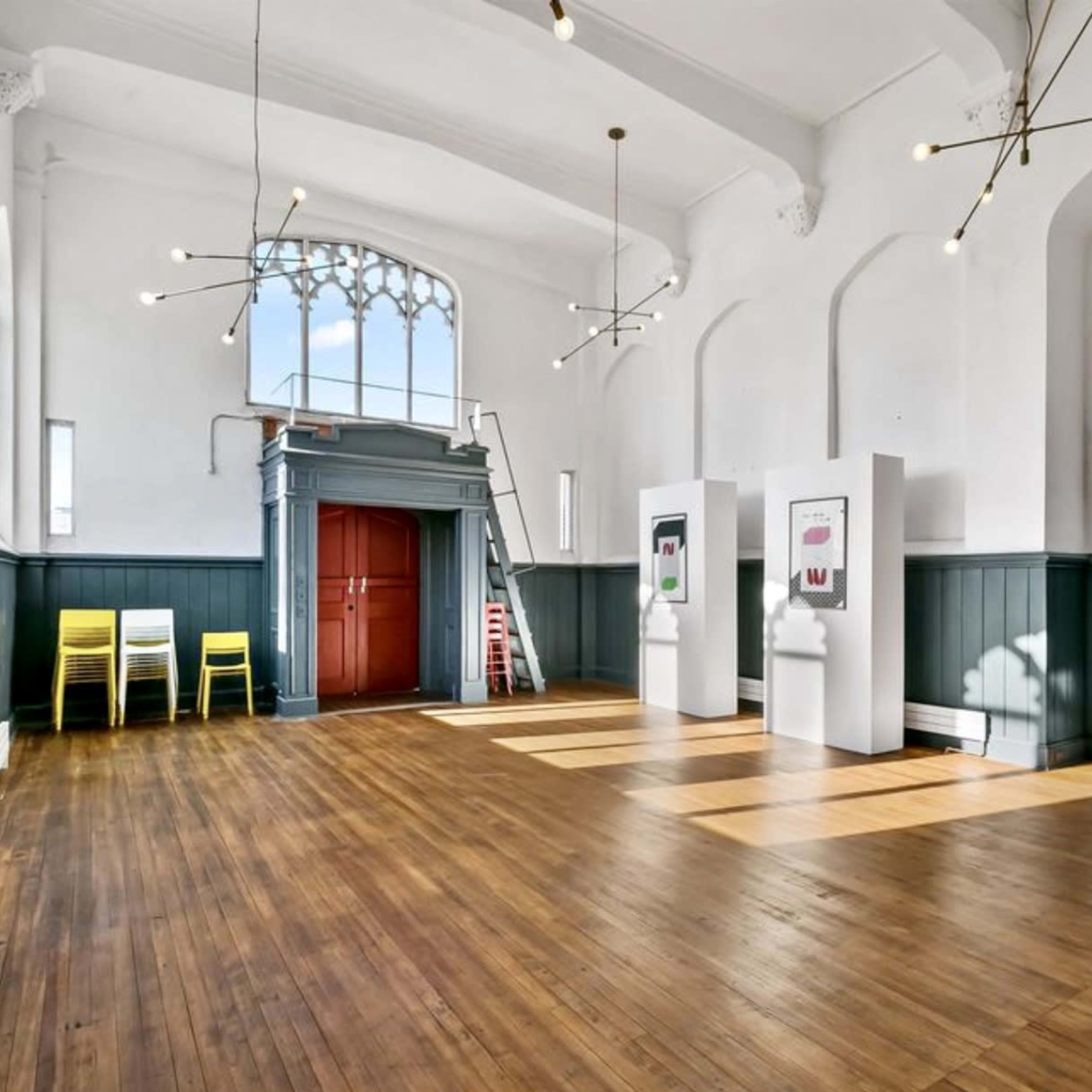 An empty room with wooden floors, bright lighting, and a staircase on one side, adorned with minimalist decor including chairs and artwork.