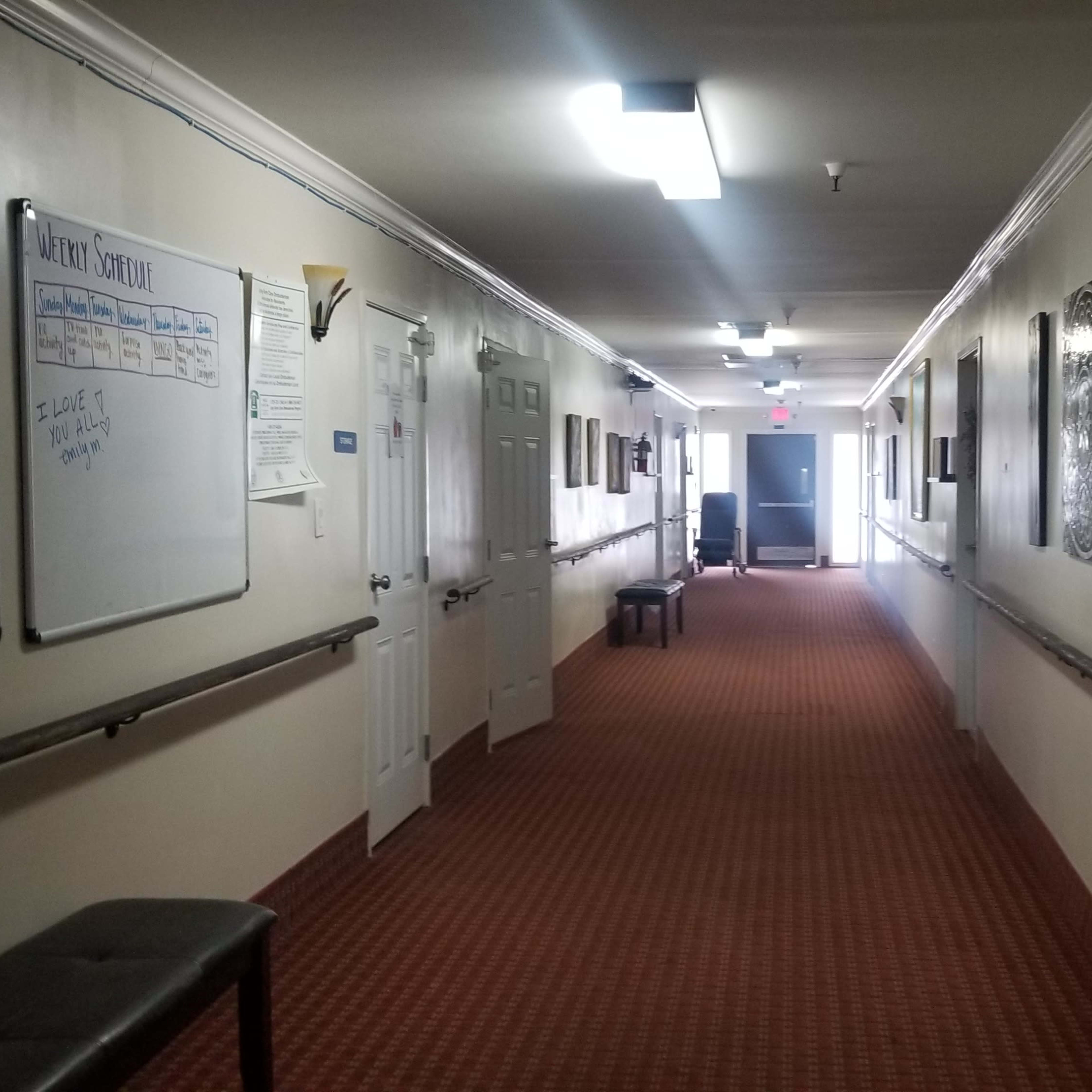 A long, narrow hallway with carpeted flooring, lined with doors and framed artwork, leads to an exit at the far end.