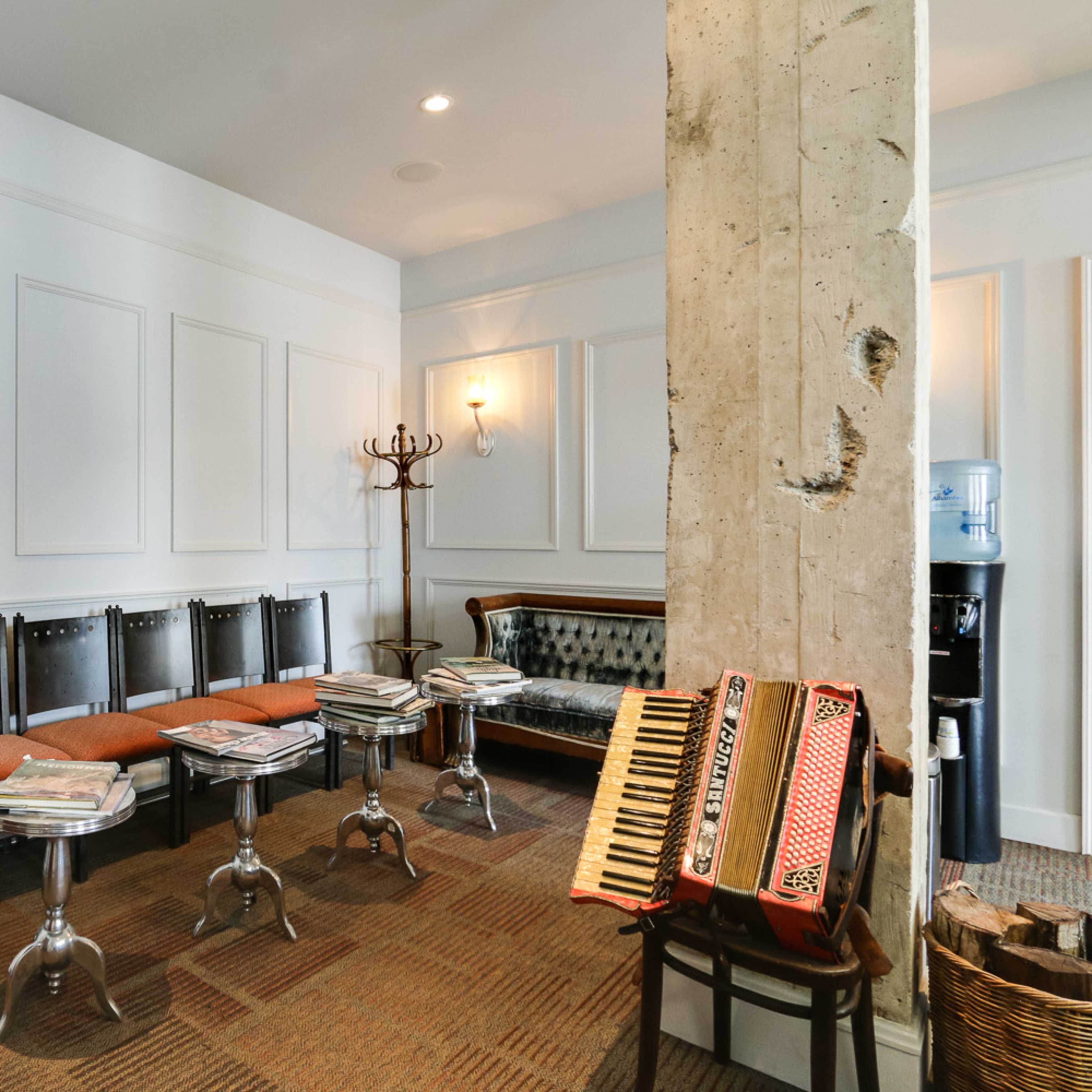 A waiting area with a row of metal and upholstered chairs, a vintage couch, a coat rack, a water cooler, and an accordion resting on a table.