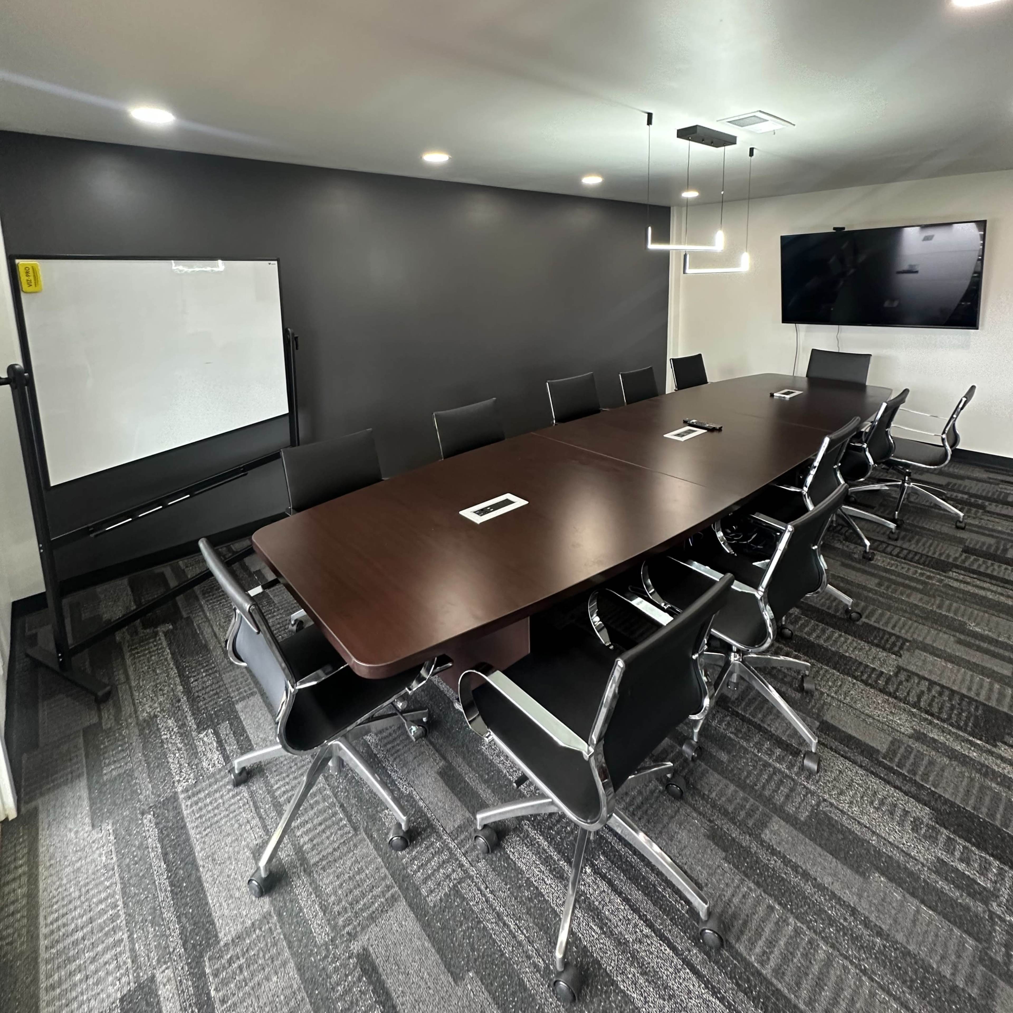 A modern conference room featuring a long wooden table surrounded by black chairs, with a whiteboard and a large screen on the walls.