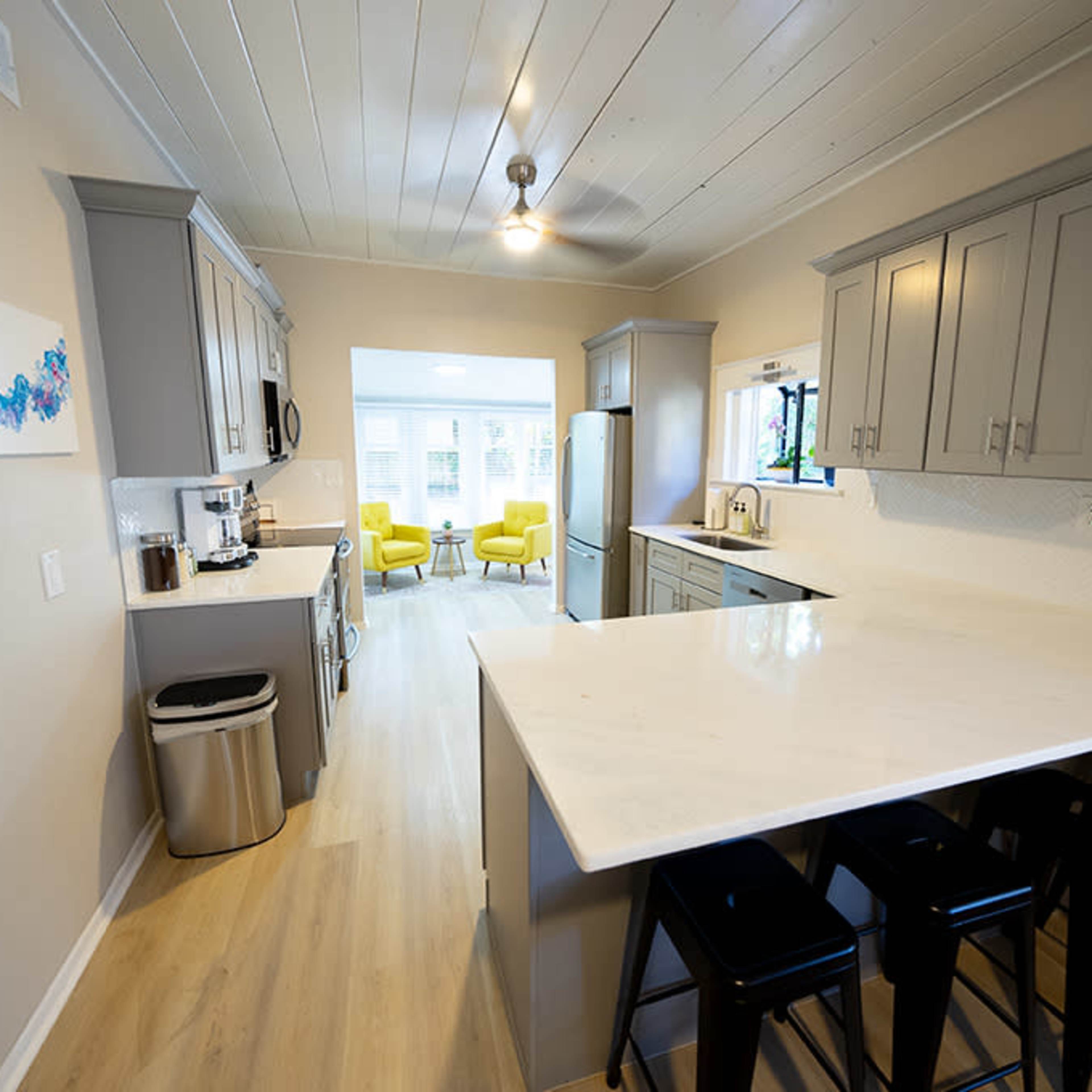 A modern kitchen with gray cabinets, a large white countertop, and a view into a living area with two yellow chairs.