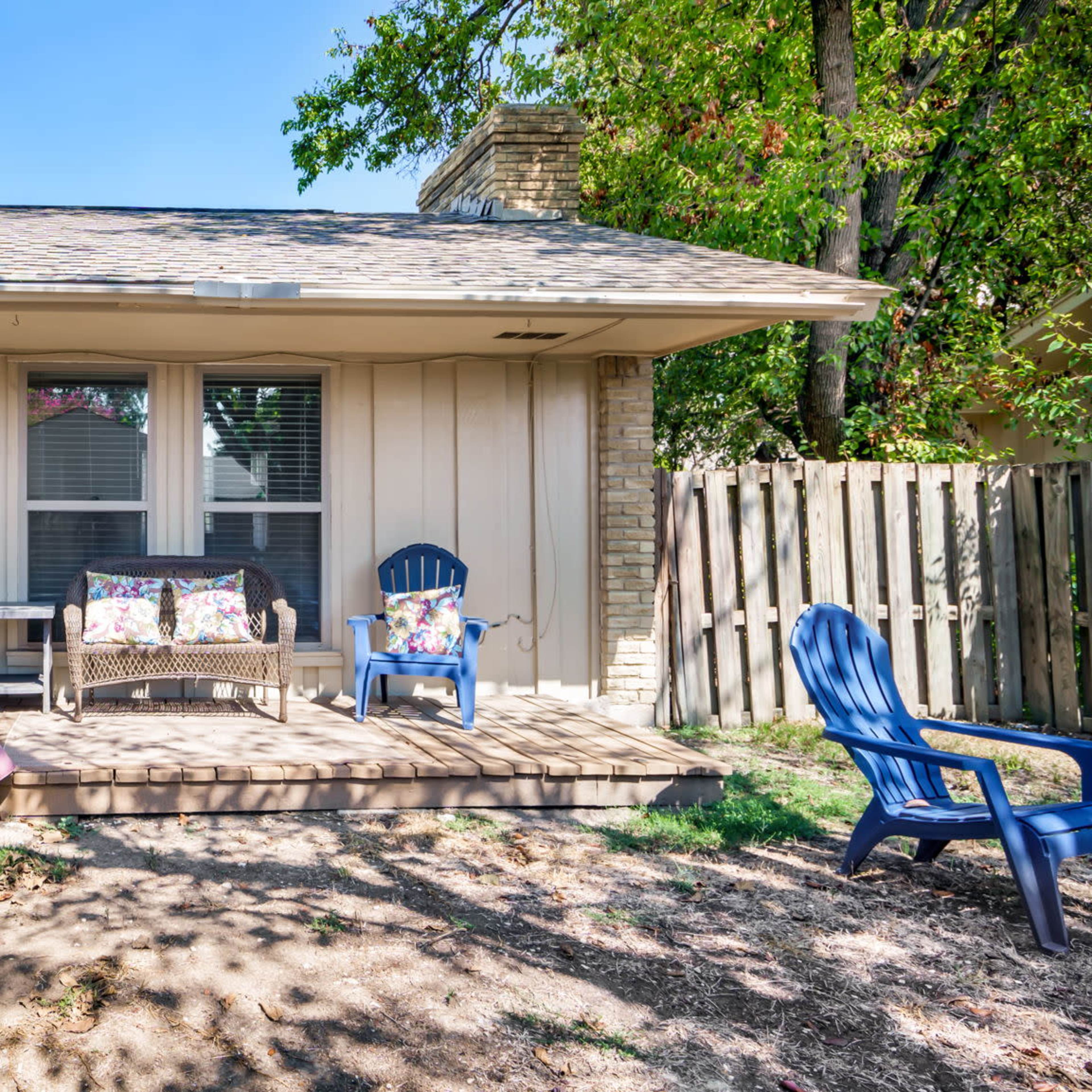 A house with a wooden deck featuring two blue chairs and a small table, surrounded by a fenced yard and trees.