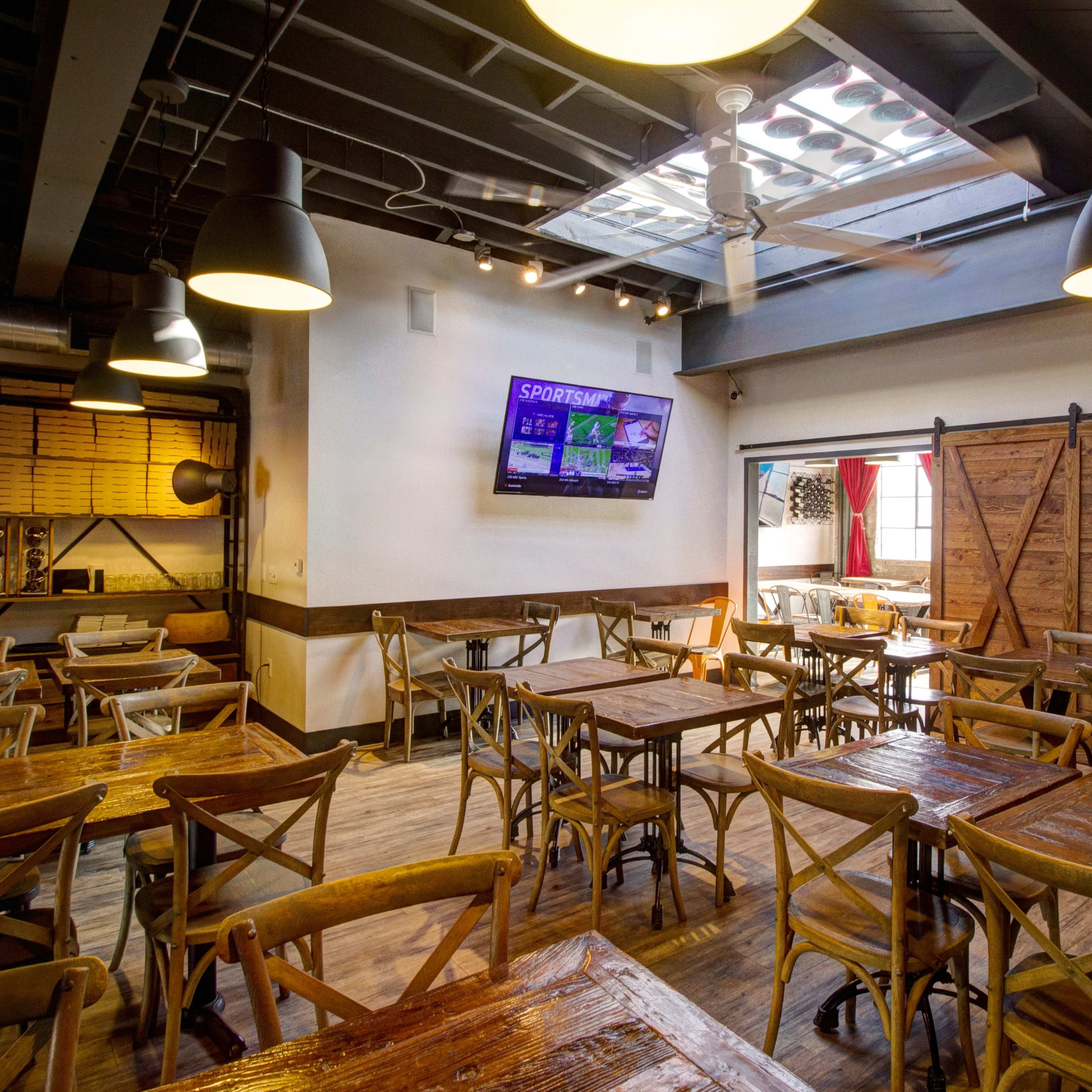 A well-lit restaurant interior with wooden tables and chairs, featuring a television mounted on the wall and a rustic wooden sliding door.